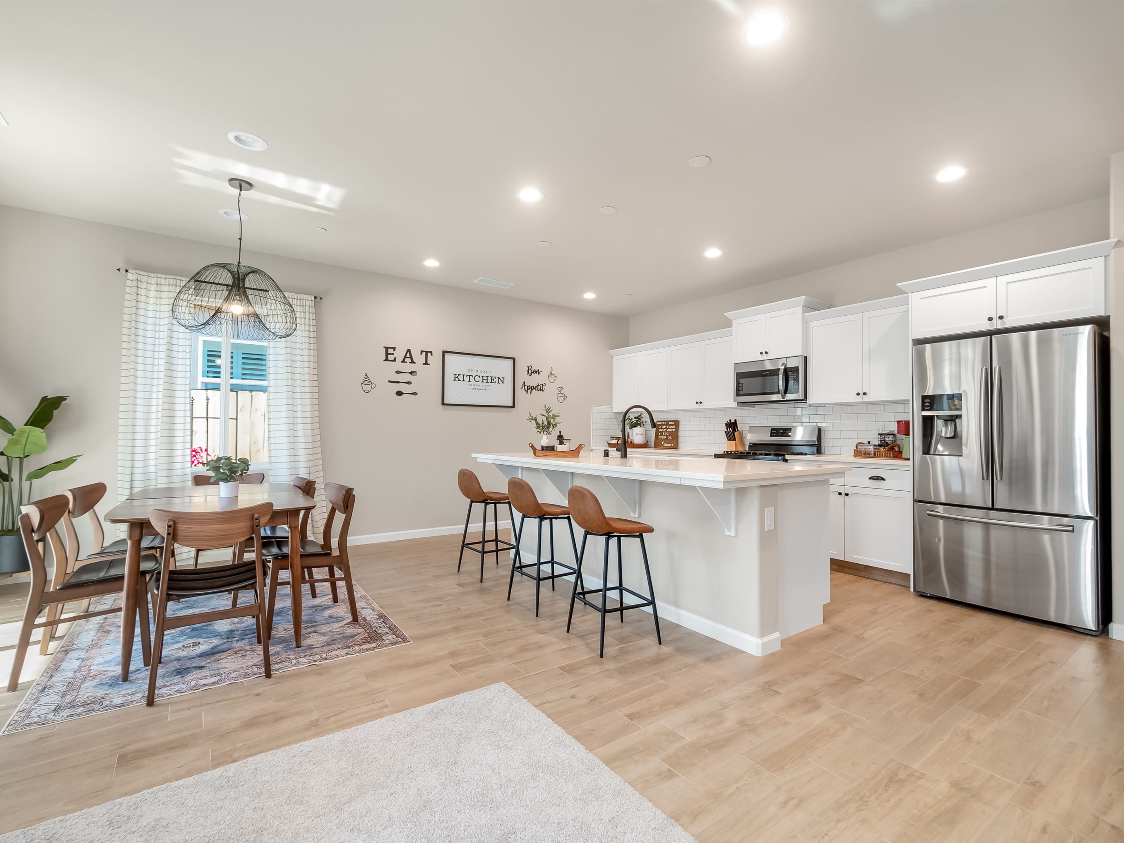 A modern kitchen and dining area features a wooden table, bar seating, stainless steel appliances, and light-colored cabinetry.