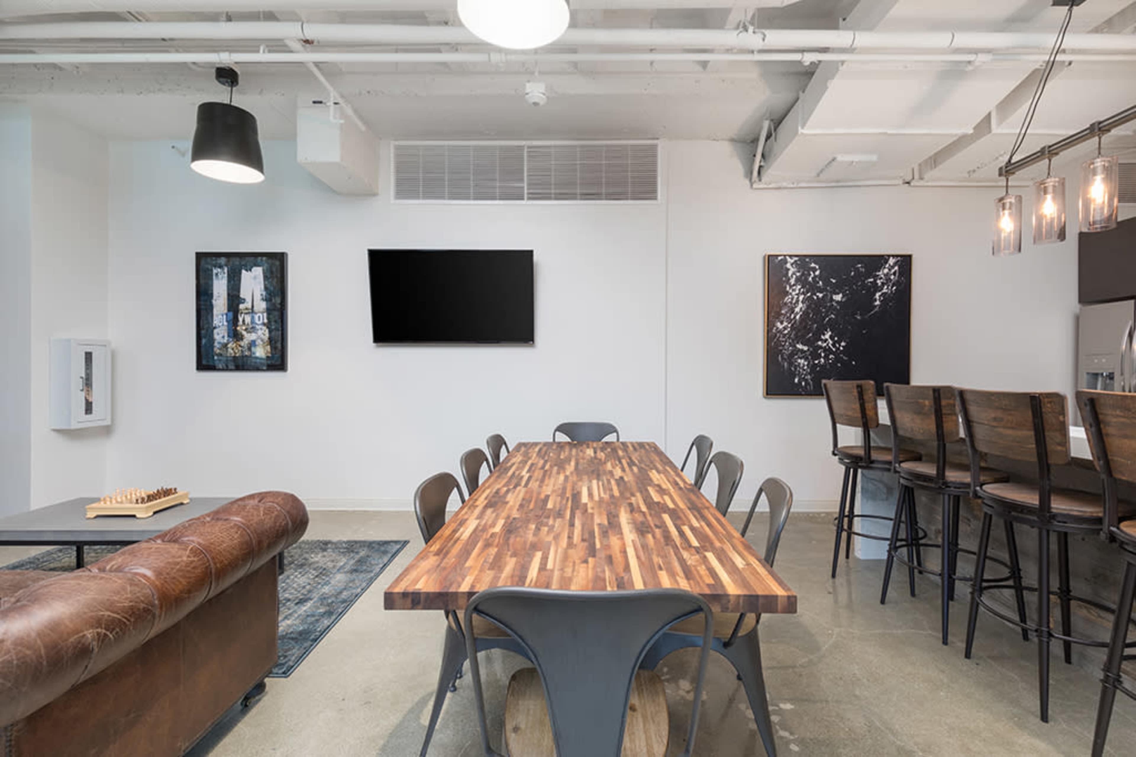 A modern meeting space features a long wooden table surrounded by metal chairs and a leather couch, with a large flat-screen TV and artworks on the walls.