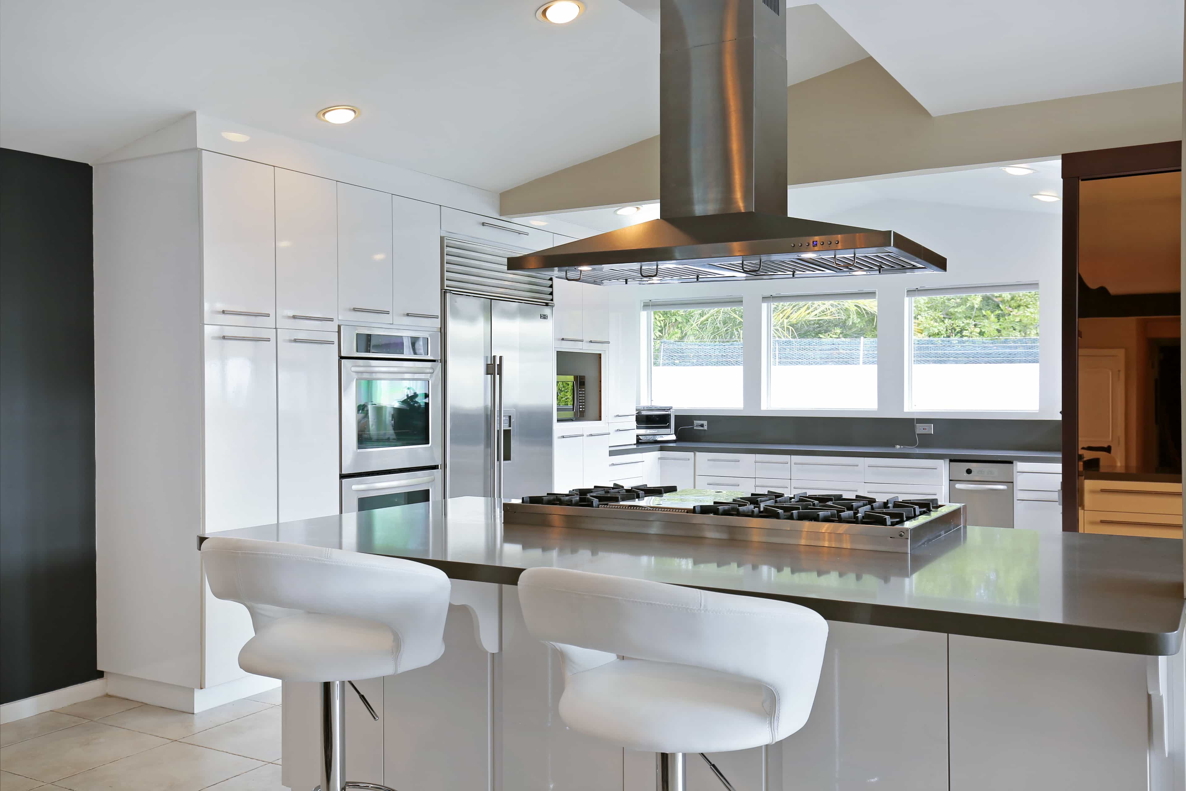 A modern kitchen features white cabinetry, a central island with a gas cooktop, and large windows providing ample natural light.