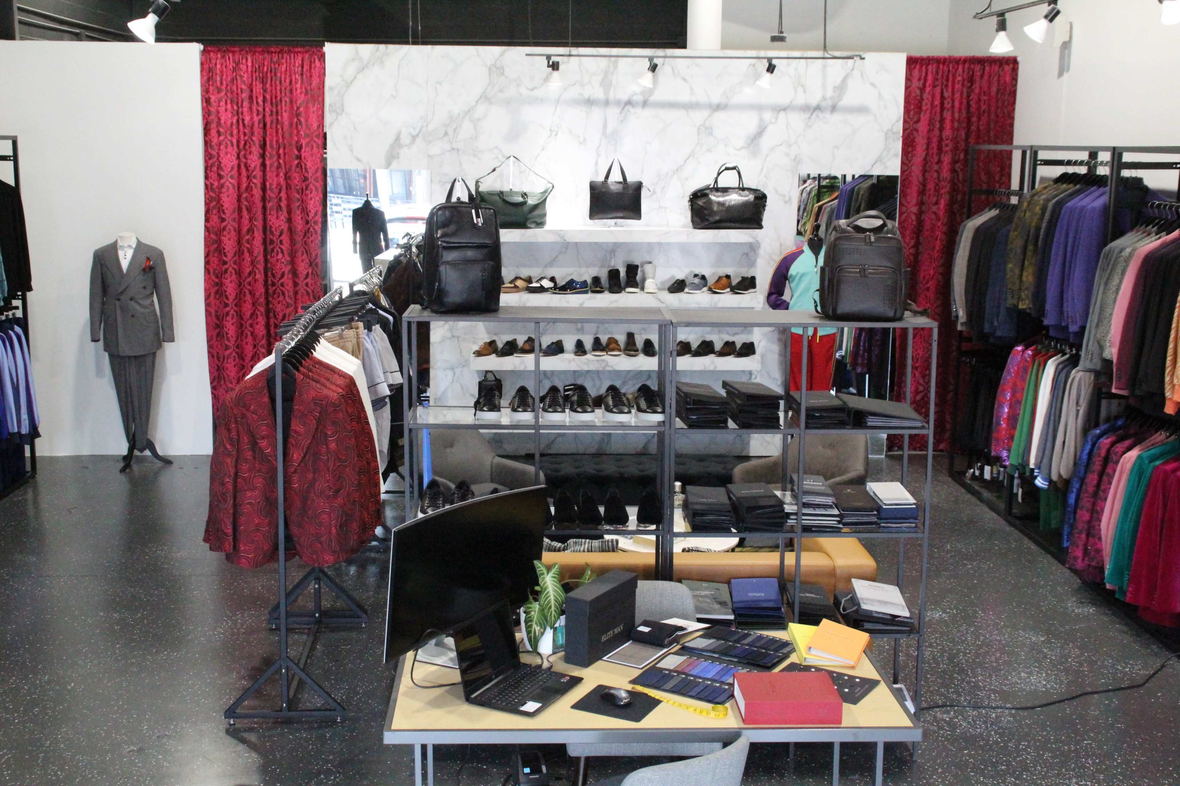 The image shows a clothing store featuring a variety of suits, bags, and shoes arranged on racks and shelves, with a desk set up in the foreground.