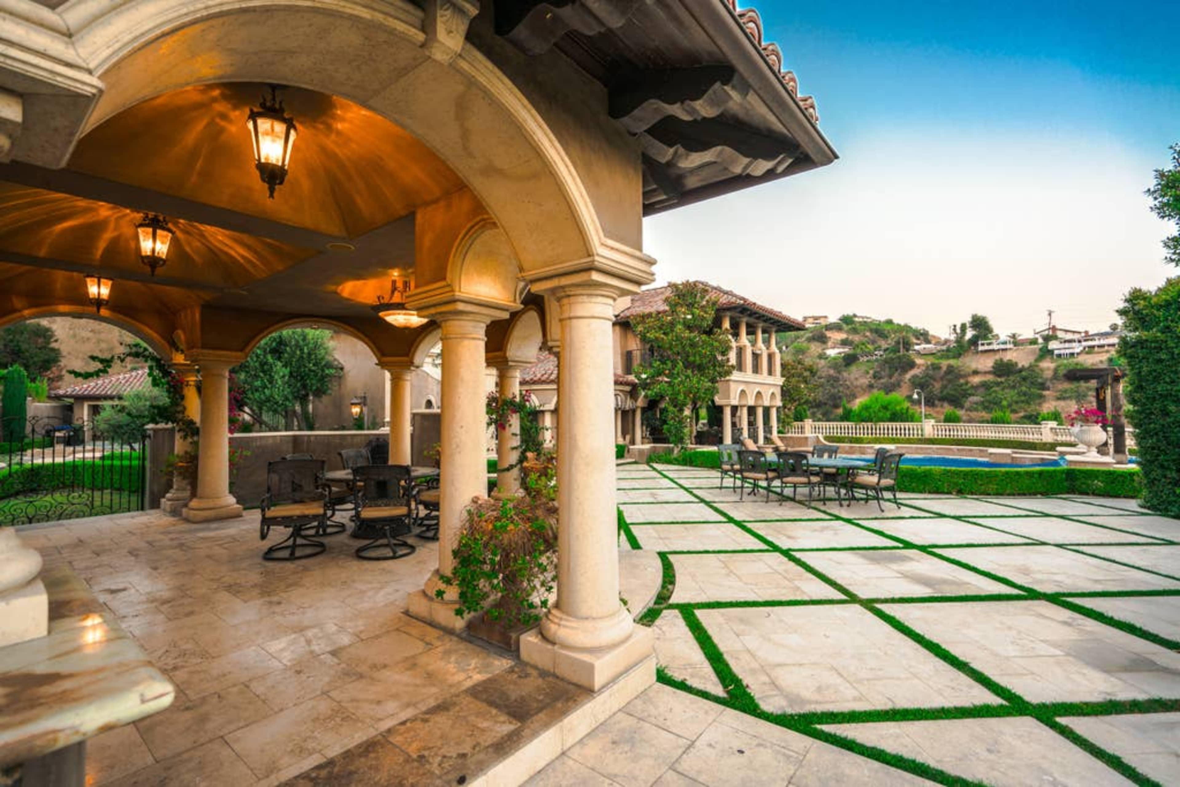 The image shows a spacious outdoor area of a luxurious estate, featuring stone columns, outdoor seating, and a landscaped garden with a pool in the background.