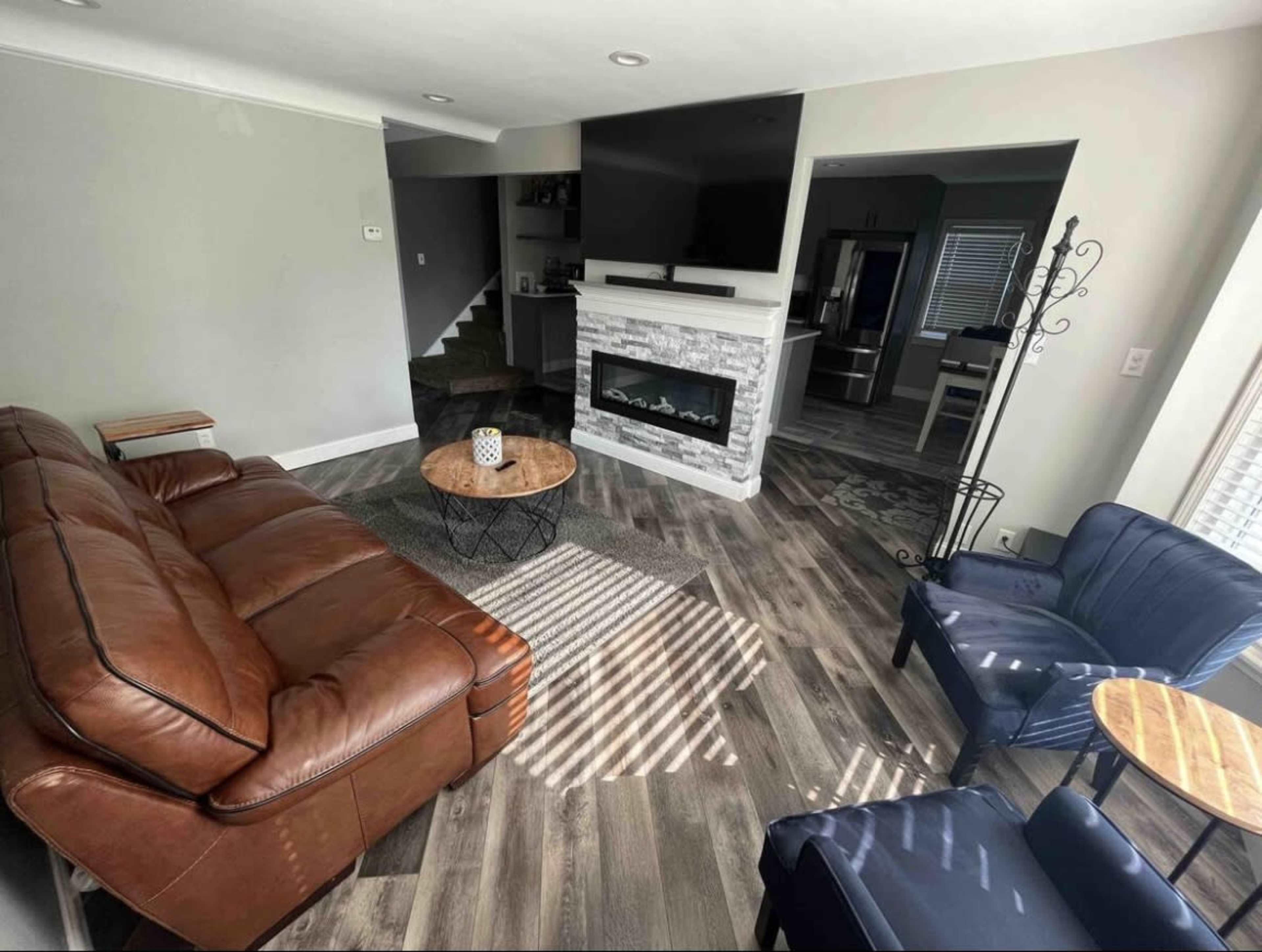 A living room with a brown leather sofa, two blue chairs, a round coffee table, and a stone fireplace beneath a television.