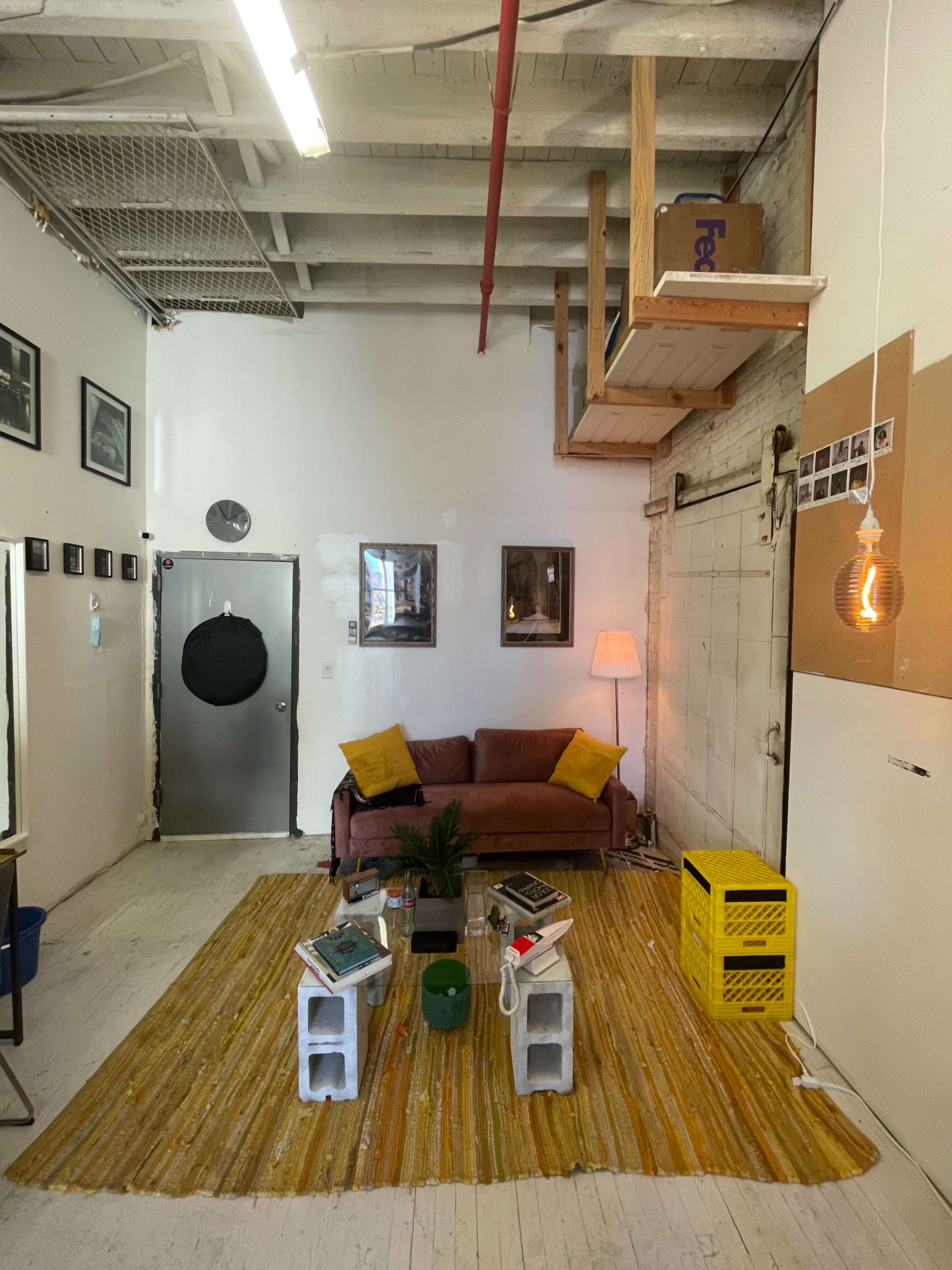 A minimalist living space with a brown couch, a yellow rug, a small table, and a lofted sleeping area, illuminated by a hanging light fixture.