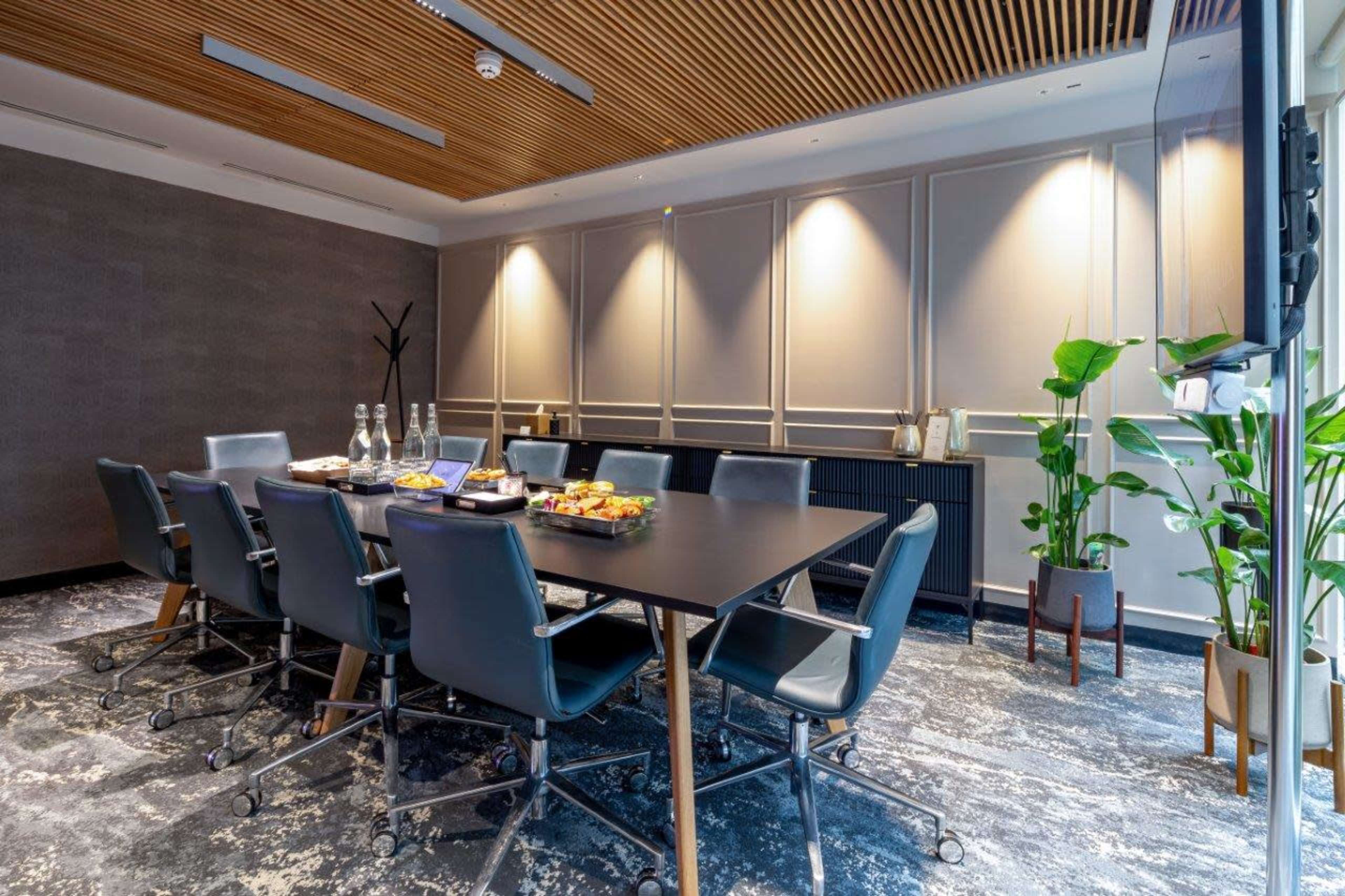 A conference room is set up with a large table surrounded by chairs, featuring refreshments and plants against a backdrop of soft lighting and textured walls.