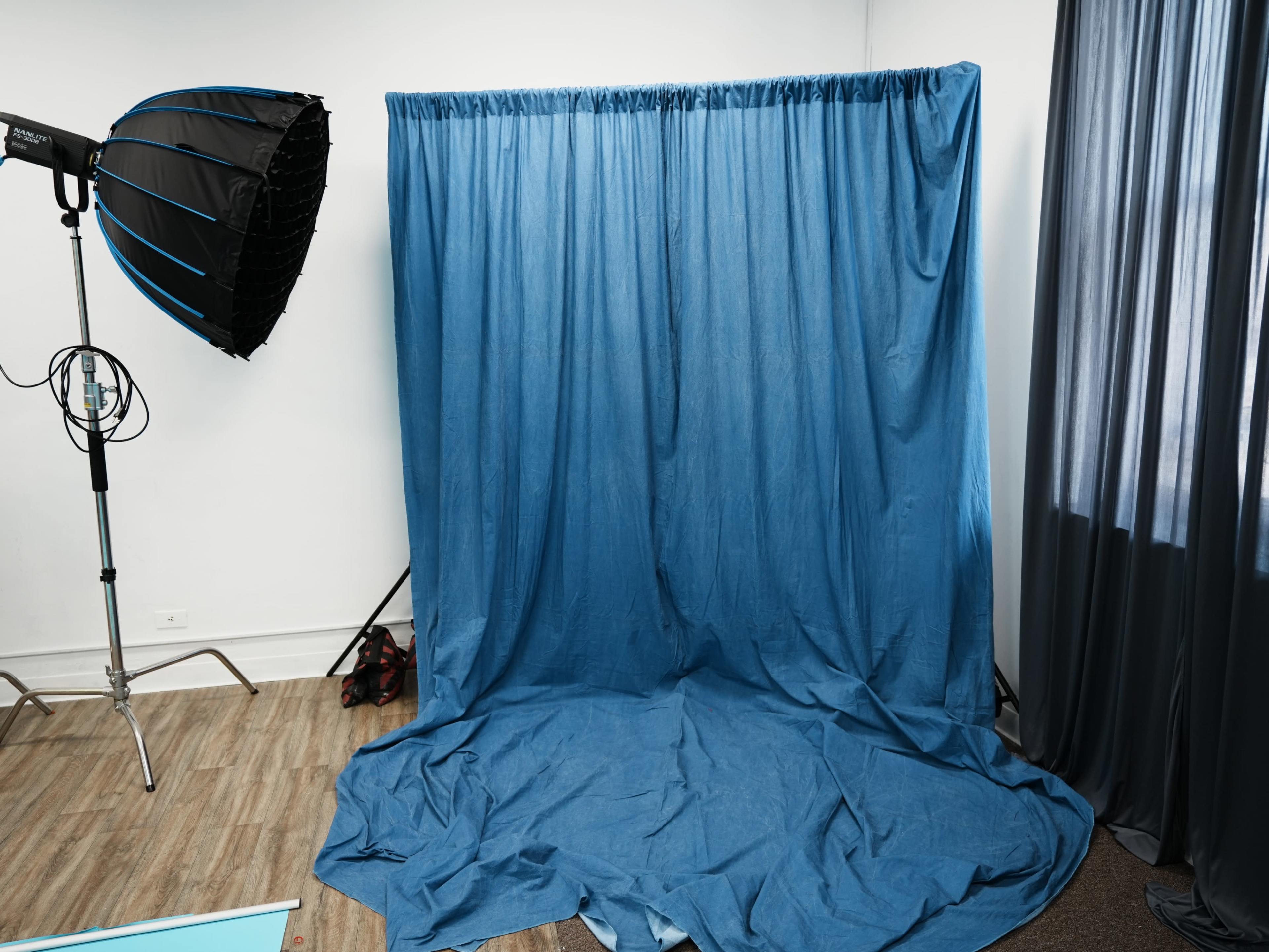 The image shows a photography studio setup with a blue backdrop draped against a wall and a large softbox light on a stand nearby.