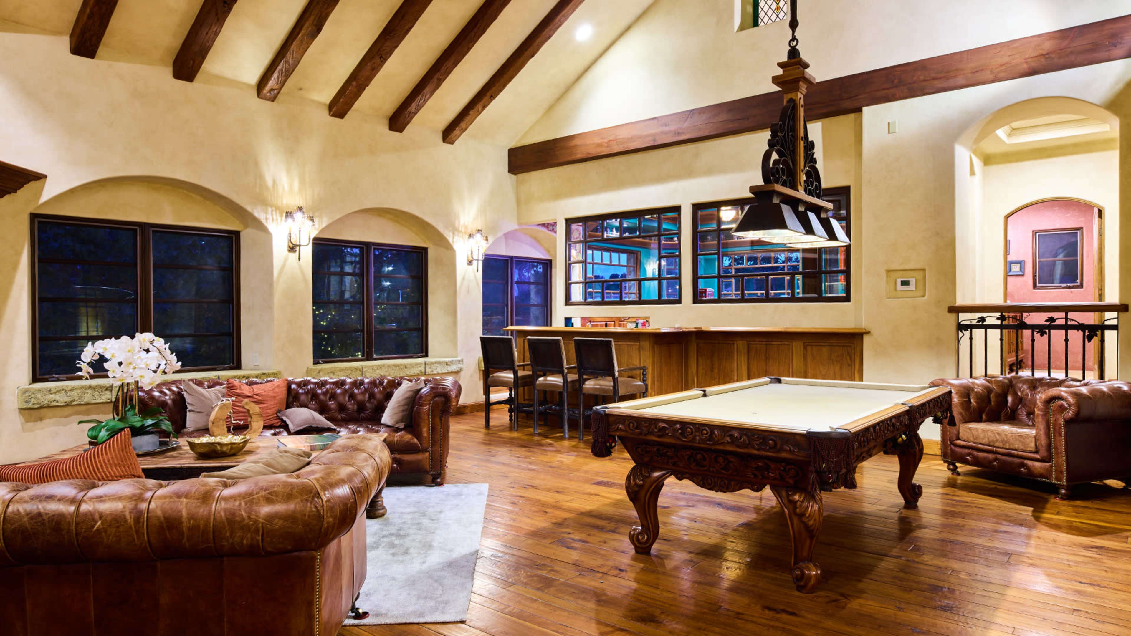 The room features a pool table beneath a hanging light fixture, surrounded by leather seating and large windows that let in natural light.