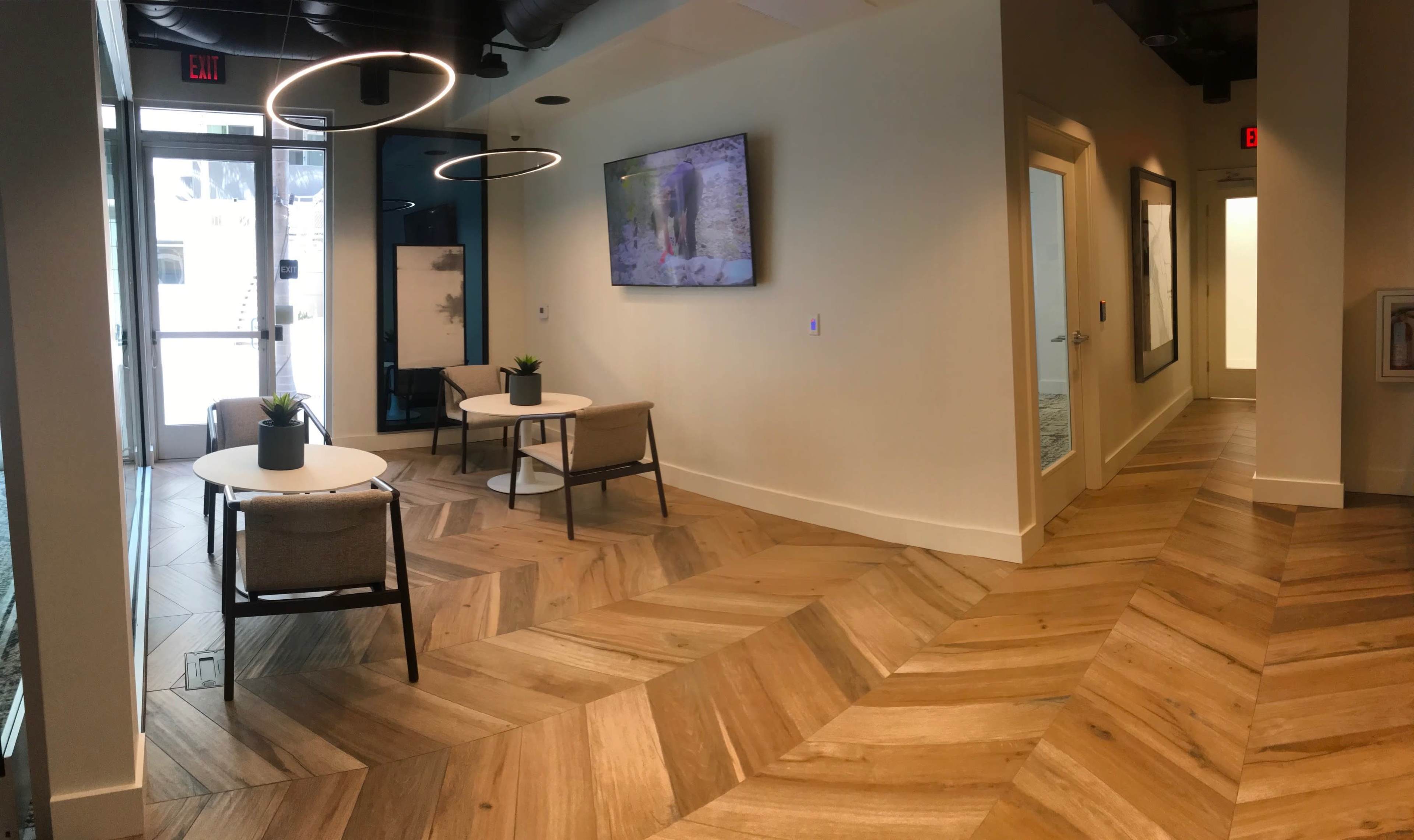 A modern open office space with a herringbone-patterned wooden floor, two small tables with plants, and a television on the wall.
