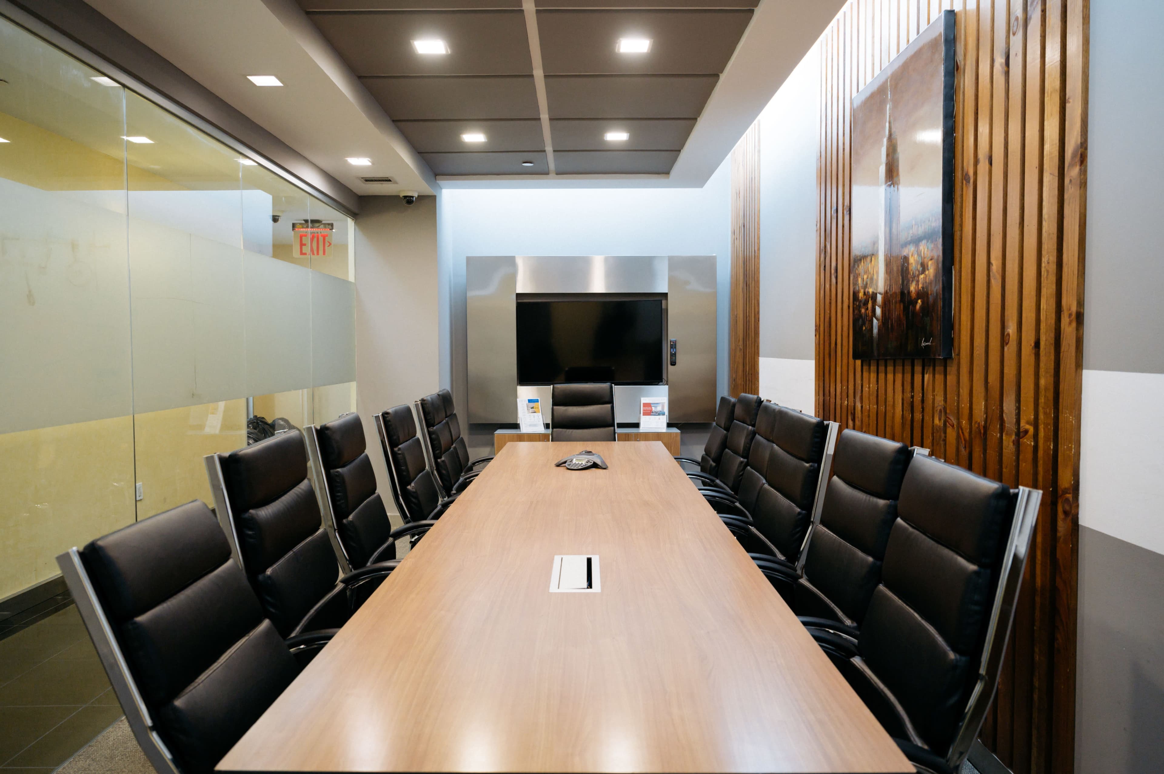 The image shows a conference room featuring a long wooden table surrounded by black leather chairs, with a television mounted on the wall and large windows allowing natural light.