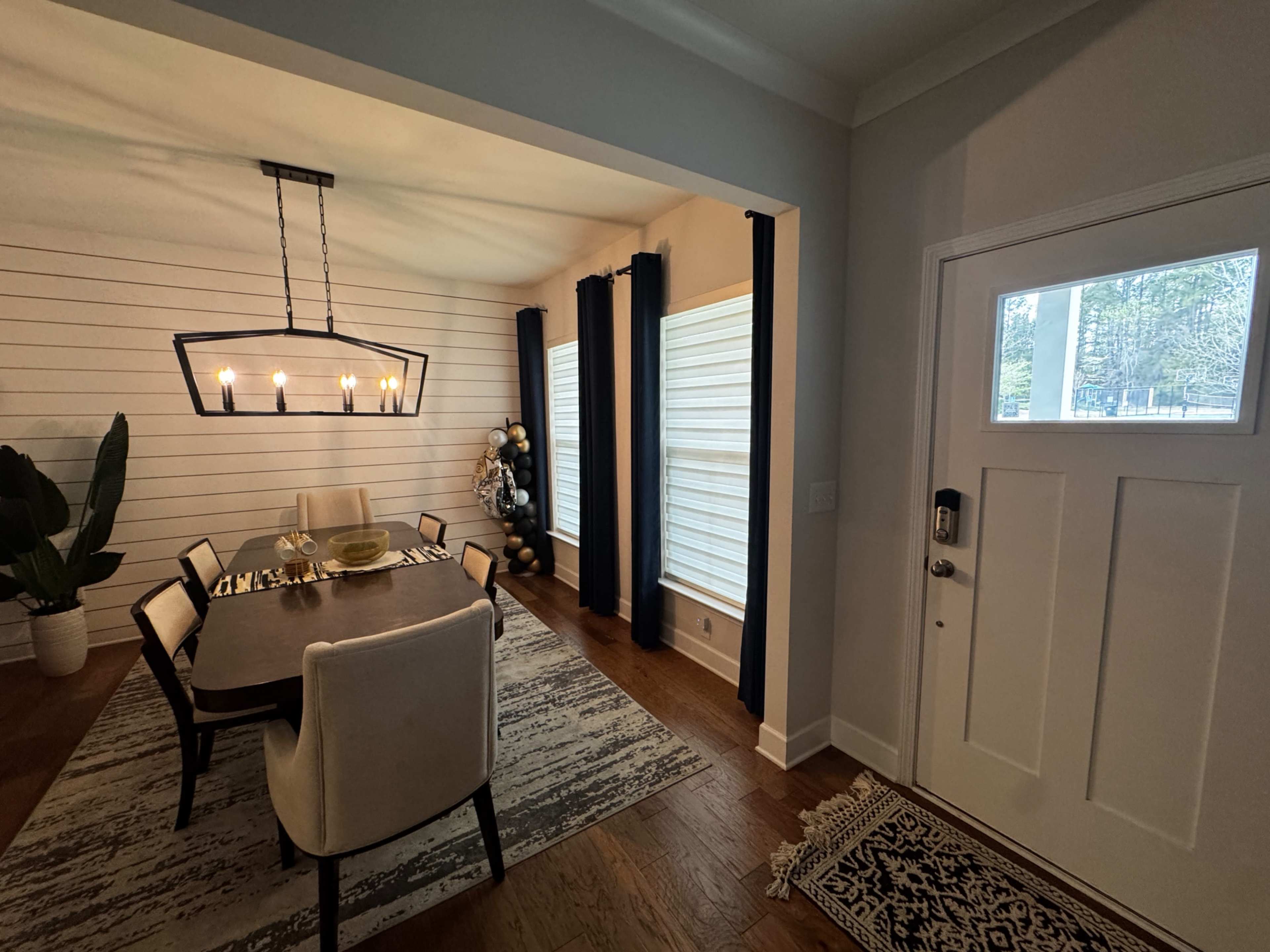 A dining area features a table with six chairs, a chandelier, and two large windows adorned with dark curtains, adjacent to an entry door.