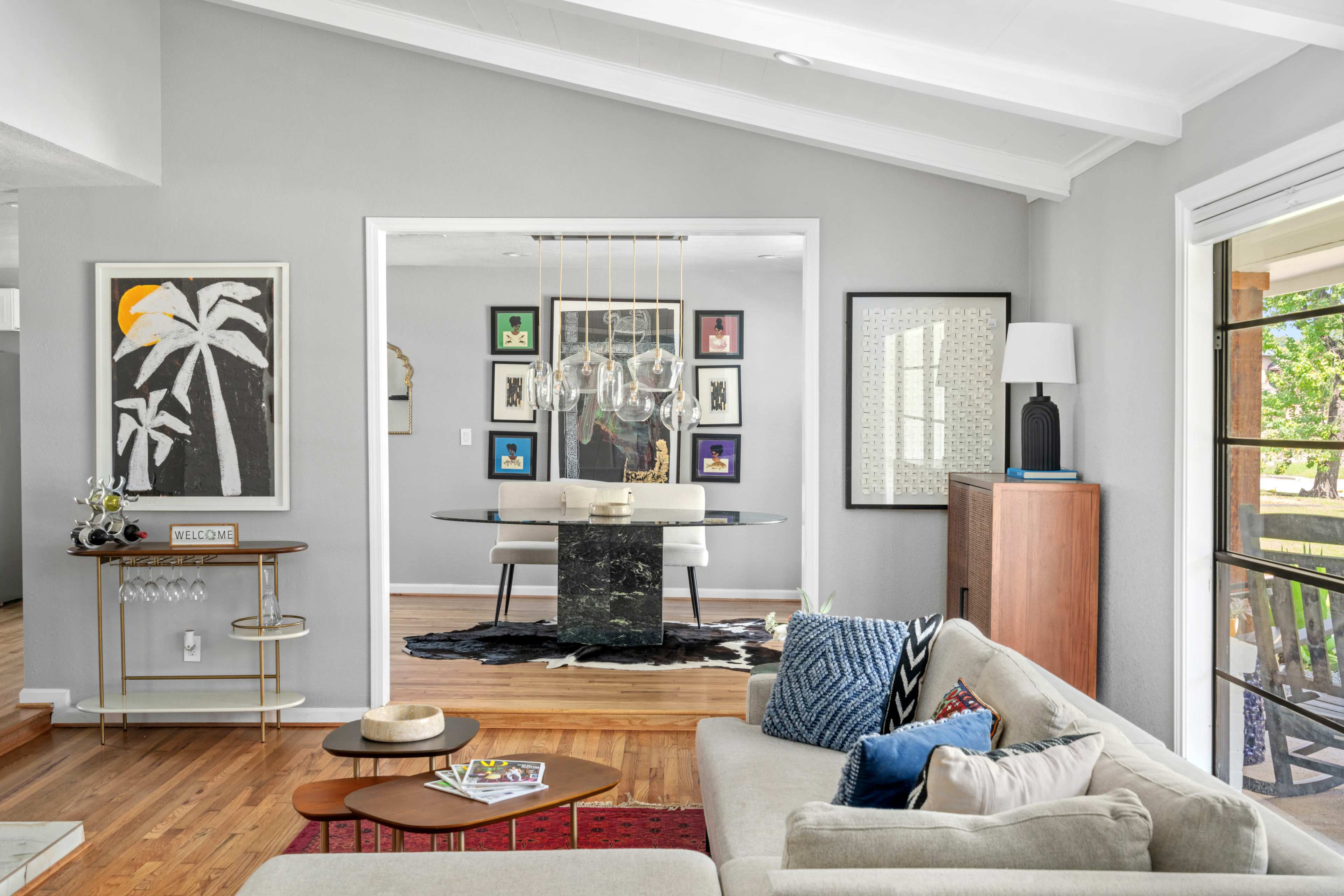 The image shows a modern living room with a gray color scheme, featuring a comfortable sofa, a coffee table, and an open view to a dining area with a black table surrounded by framed artwork.