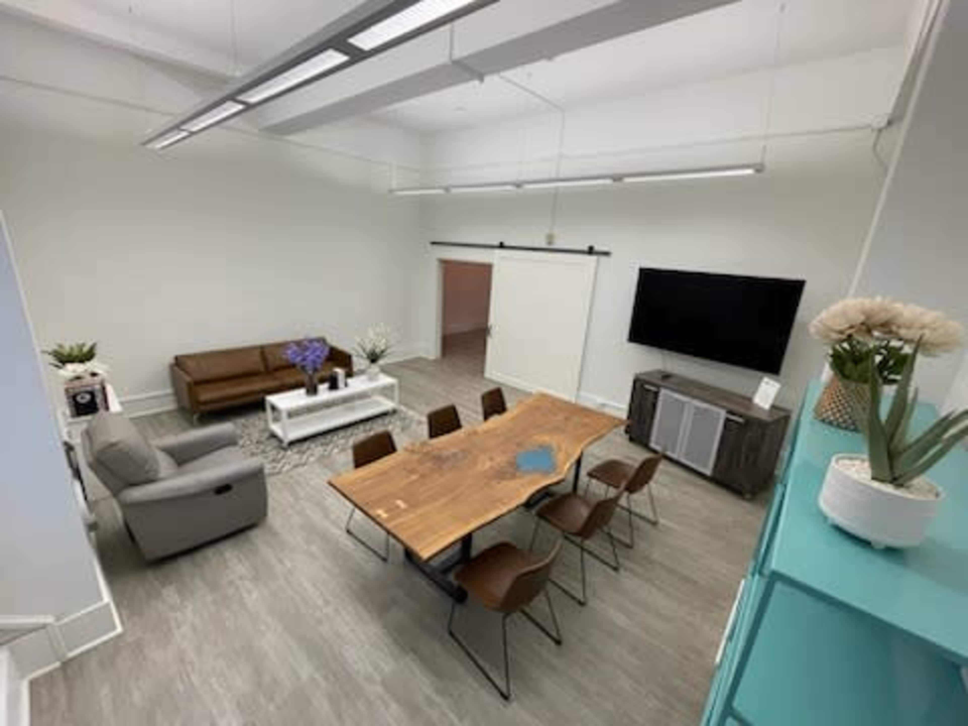 The image shows a bright, spacious room with a long wooden table surrounded by chairs, a sofa, and a television mounted on the wall.