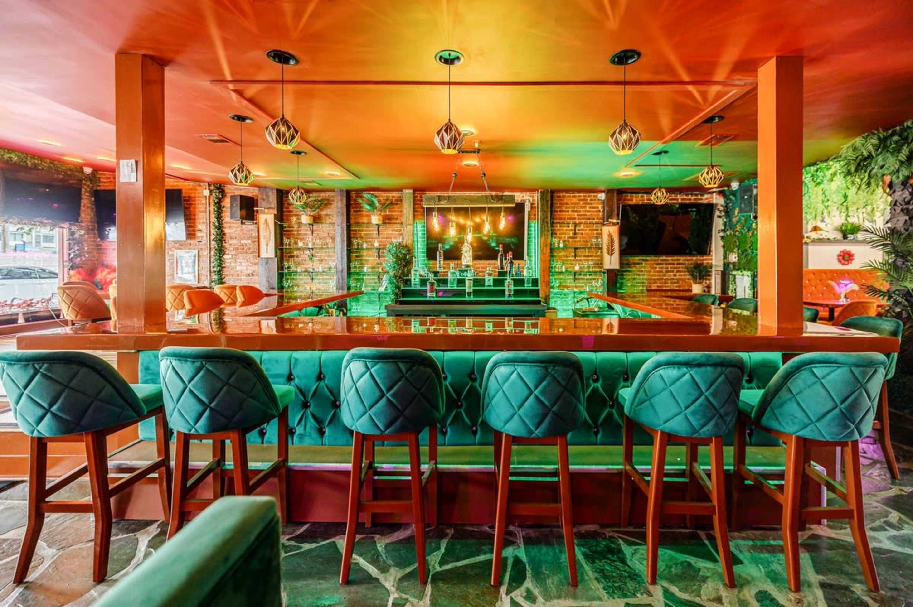 A bar area with teal upholstered seating, wooden stools, and vibrant lighting in a brick-walled establishment.