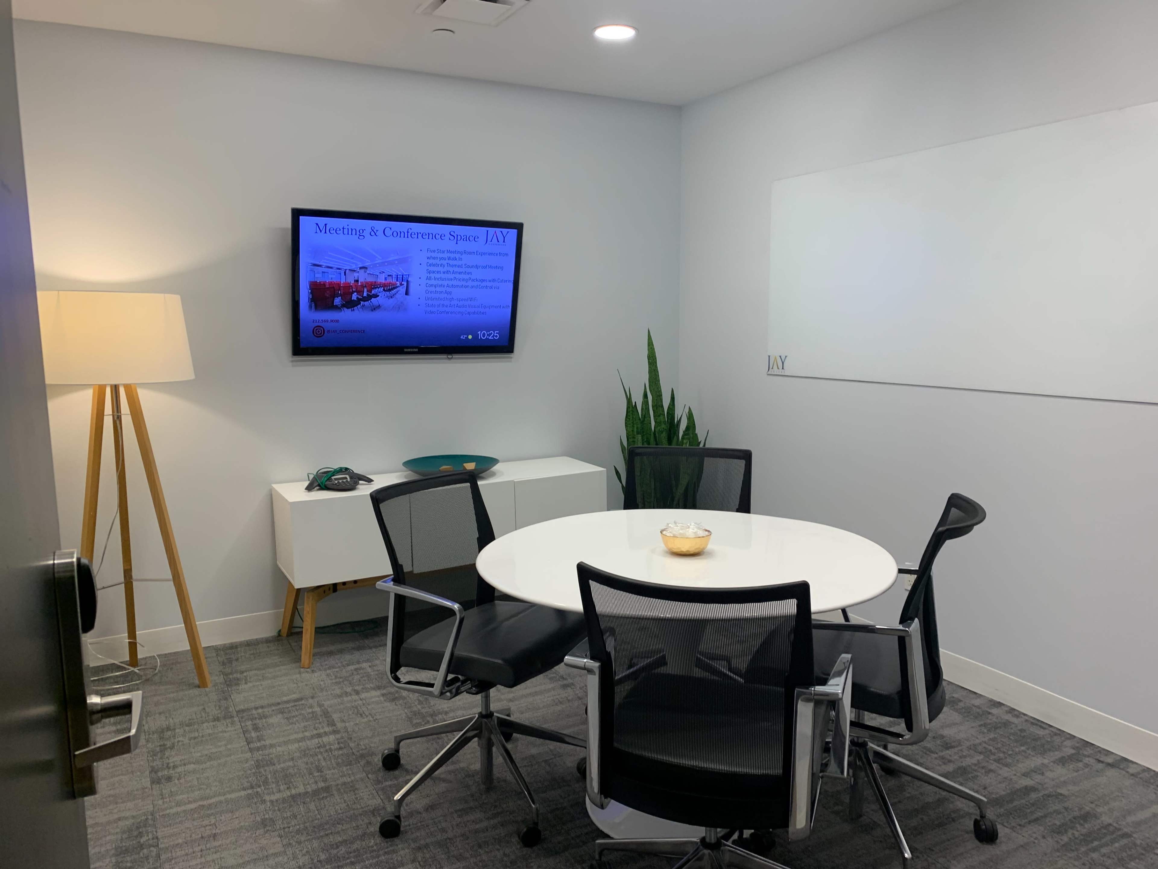 A modern conference room features a round table with four chairs, a television displaying meeting information, and a potted plant in the corner.