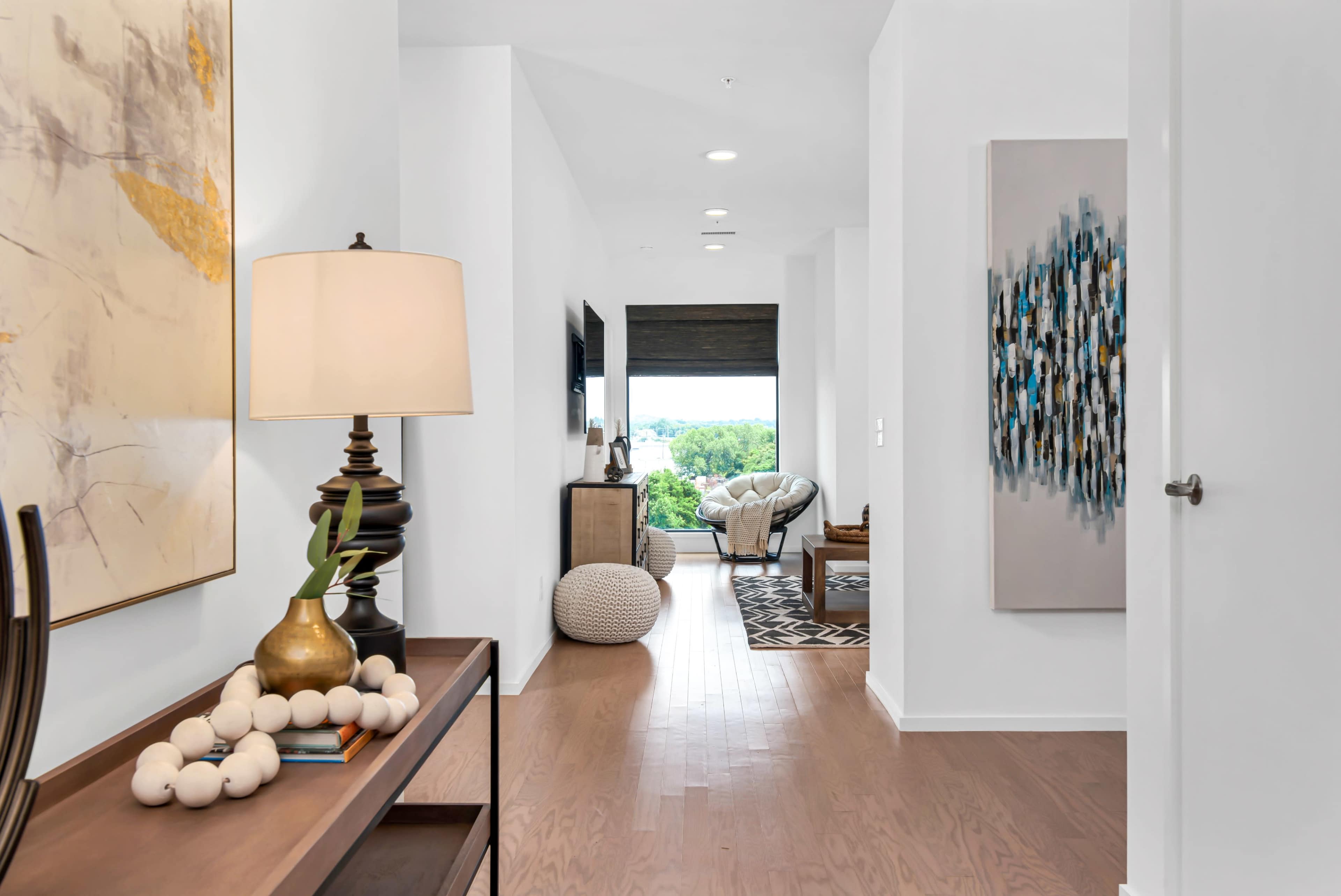 A bright hallway leading to a living space, featuring a console table with a lamp, artwork on the walls, and a window with a view of greenery.