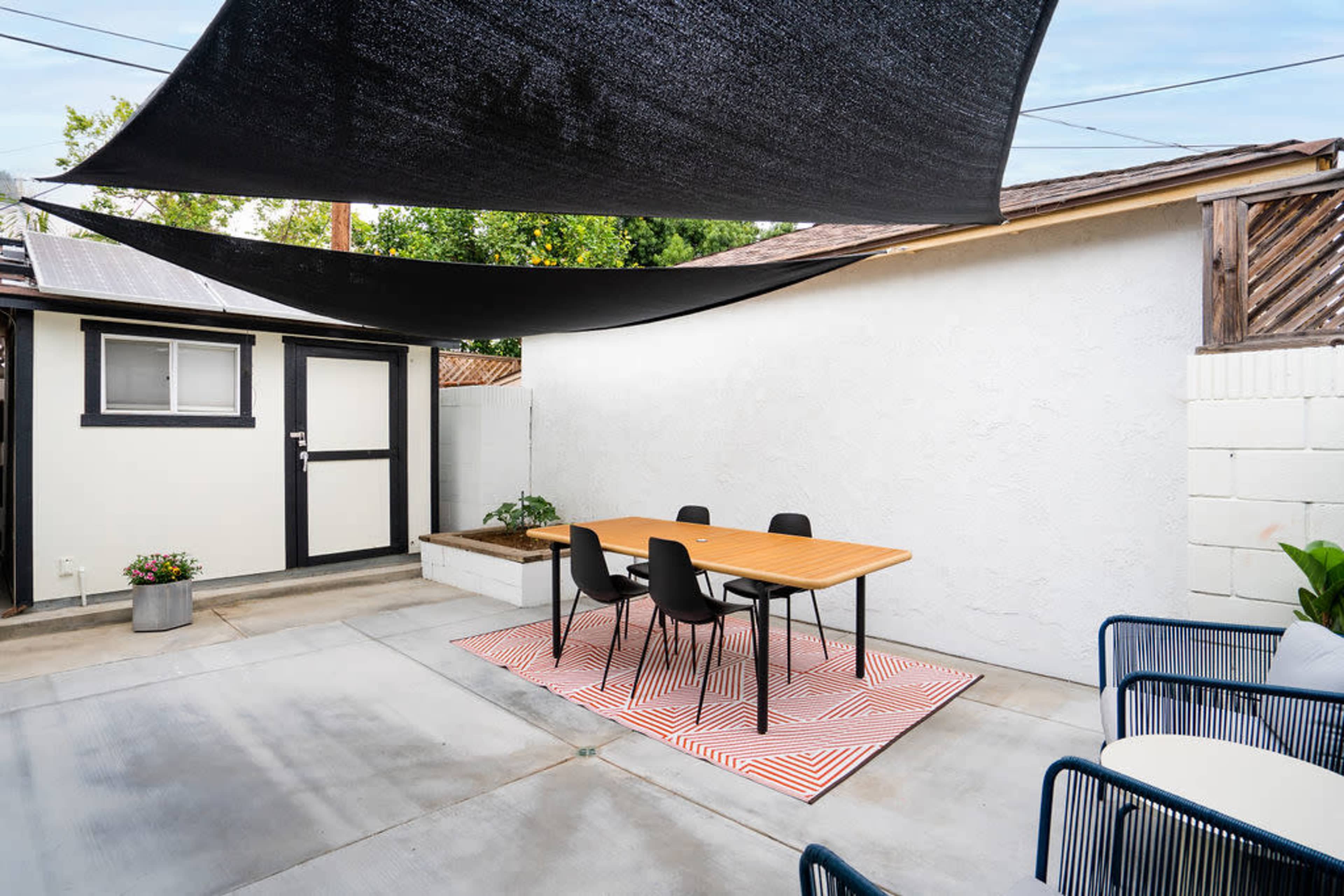 A minimalist outdoor dining area with a wooden table and black chairs, sheltered by black shade sails, adjacent to a white wall.