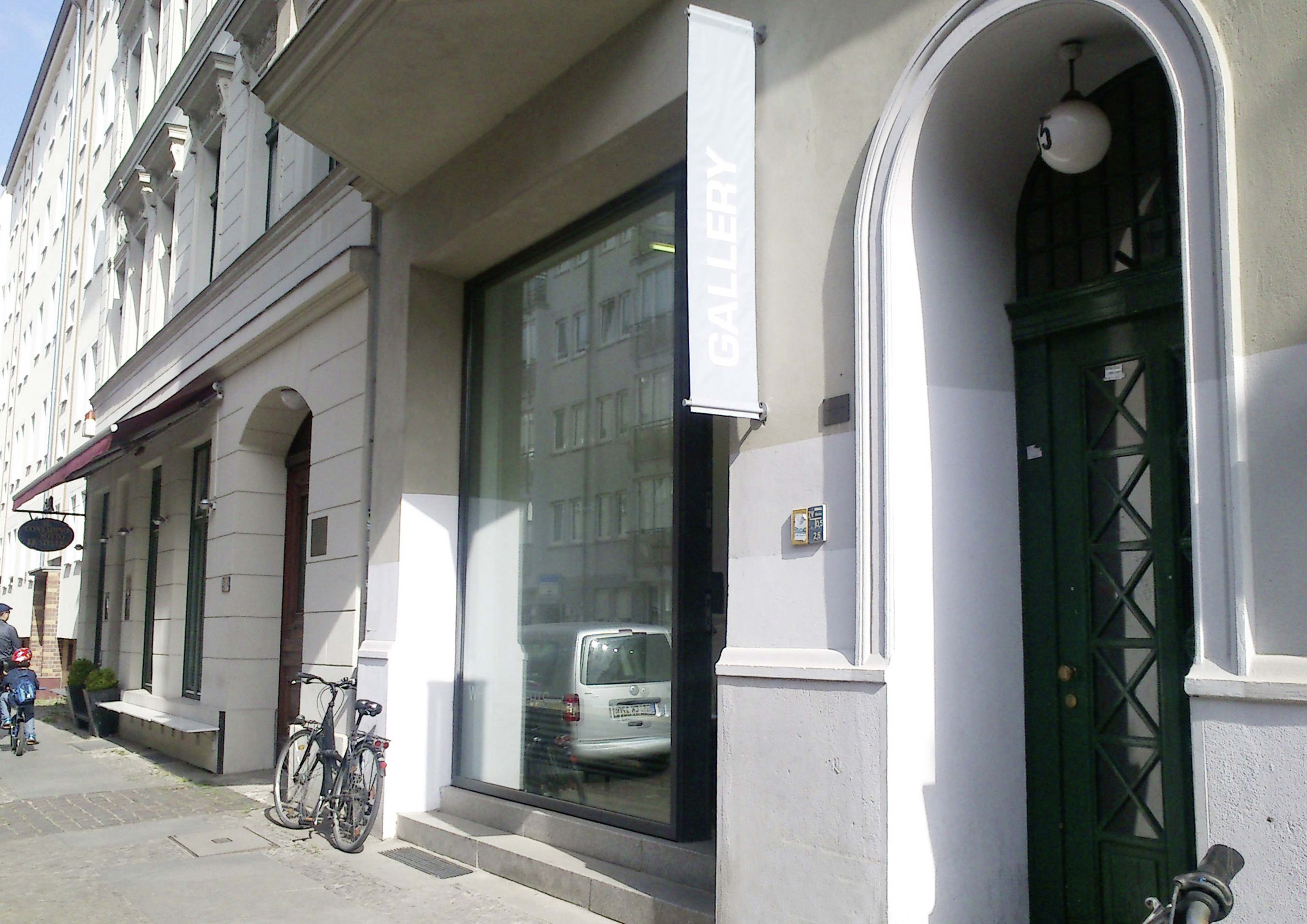 The image shows a street scene featuring a contemporary art gallery with large glass windows, a sign stating "GALLERY," and bicycles parked outside.