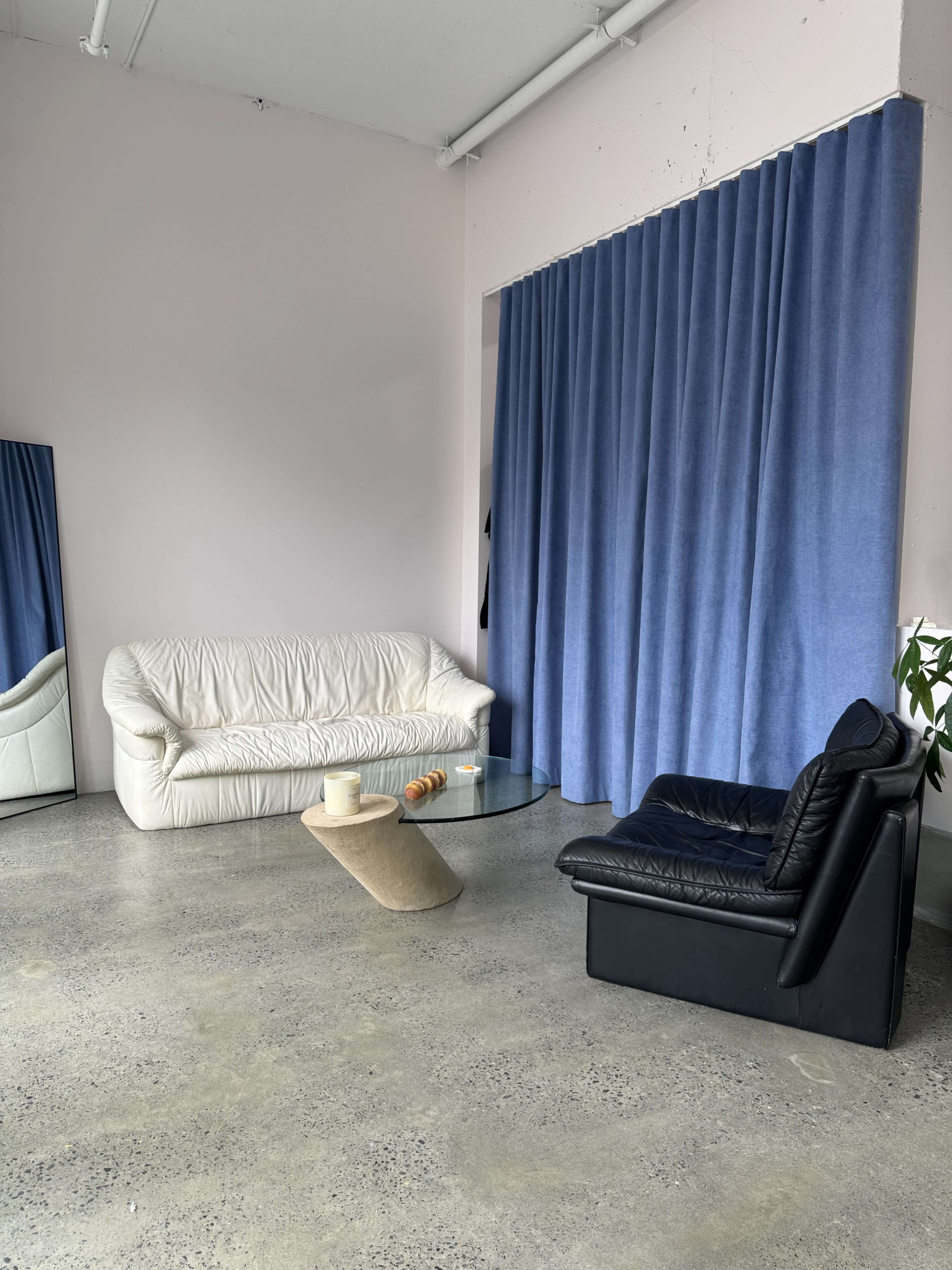 The image shows a minimalist living space with a white sofa, a black armchair, a glass coffee table, and blue curtains against a light pink wall.