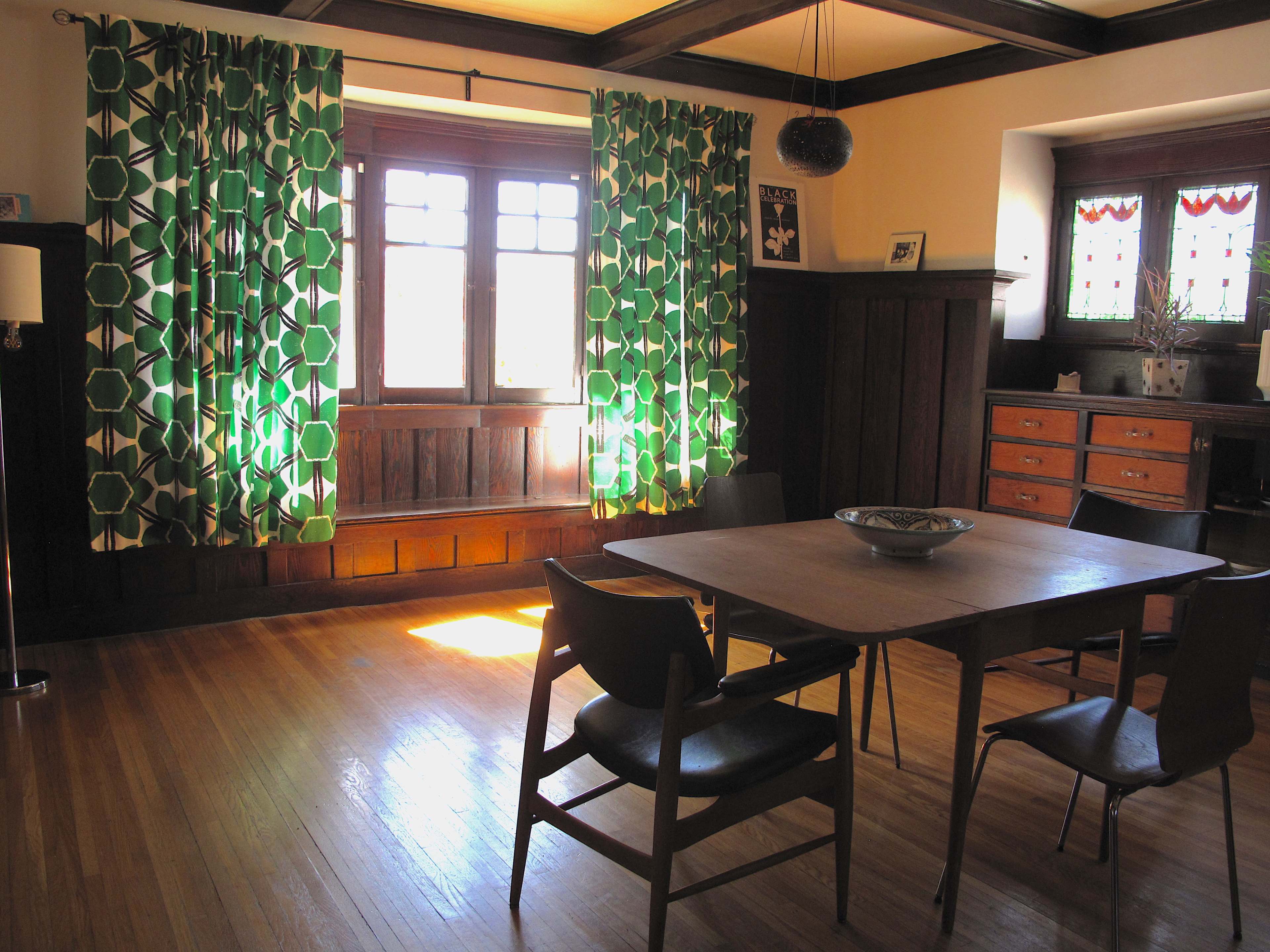 A dining area with a wooden table surrounded by chairs, featuring large green patterned curtains and a sideboard against a wooden wall.