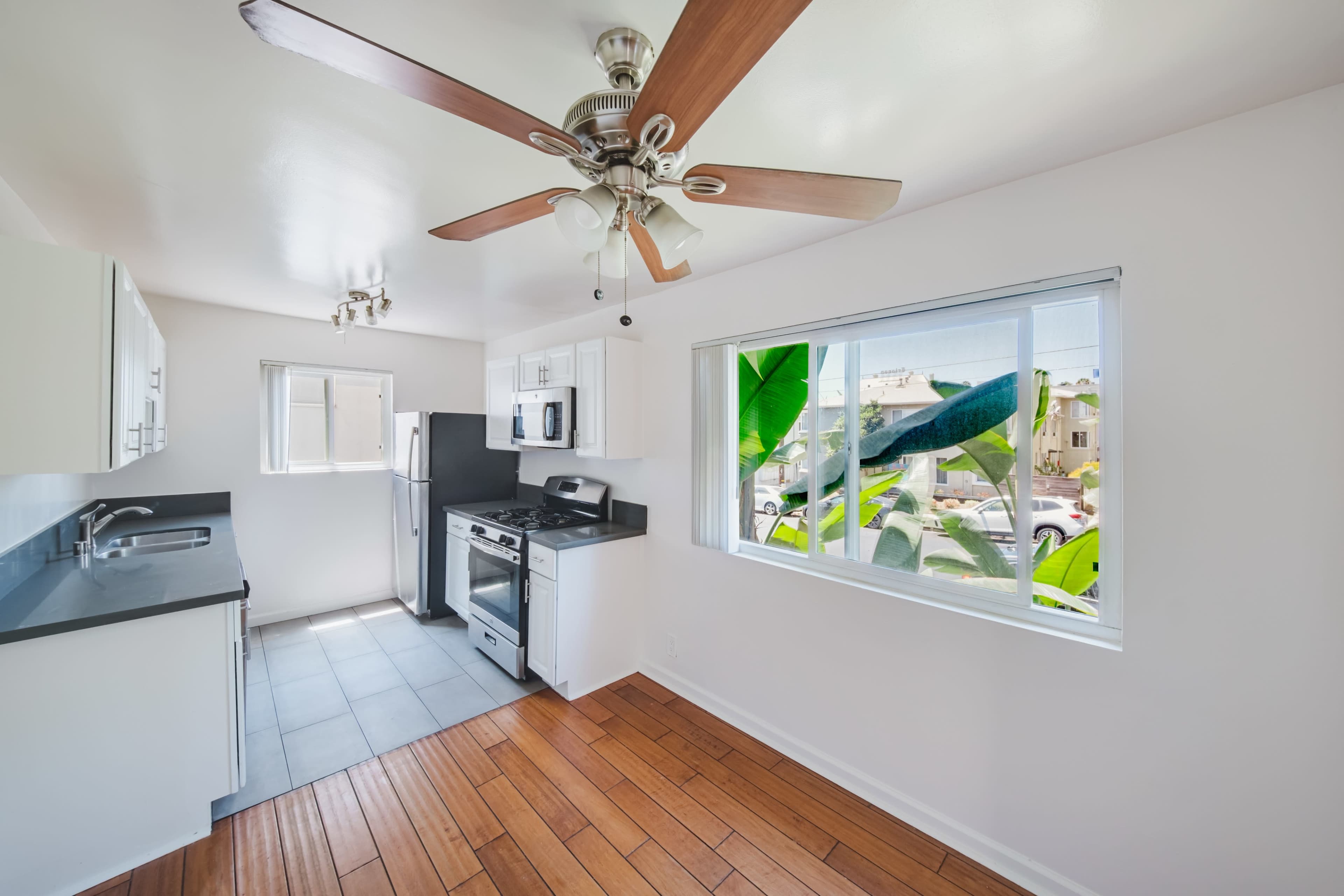 The image shows a modern kitchen with a ceiling fan, white cabinetry, stainless steel appliances, and a window overlooking a street with greenery.