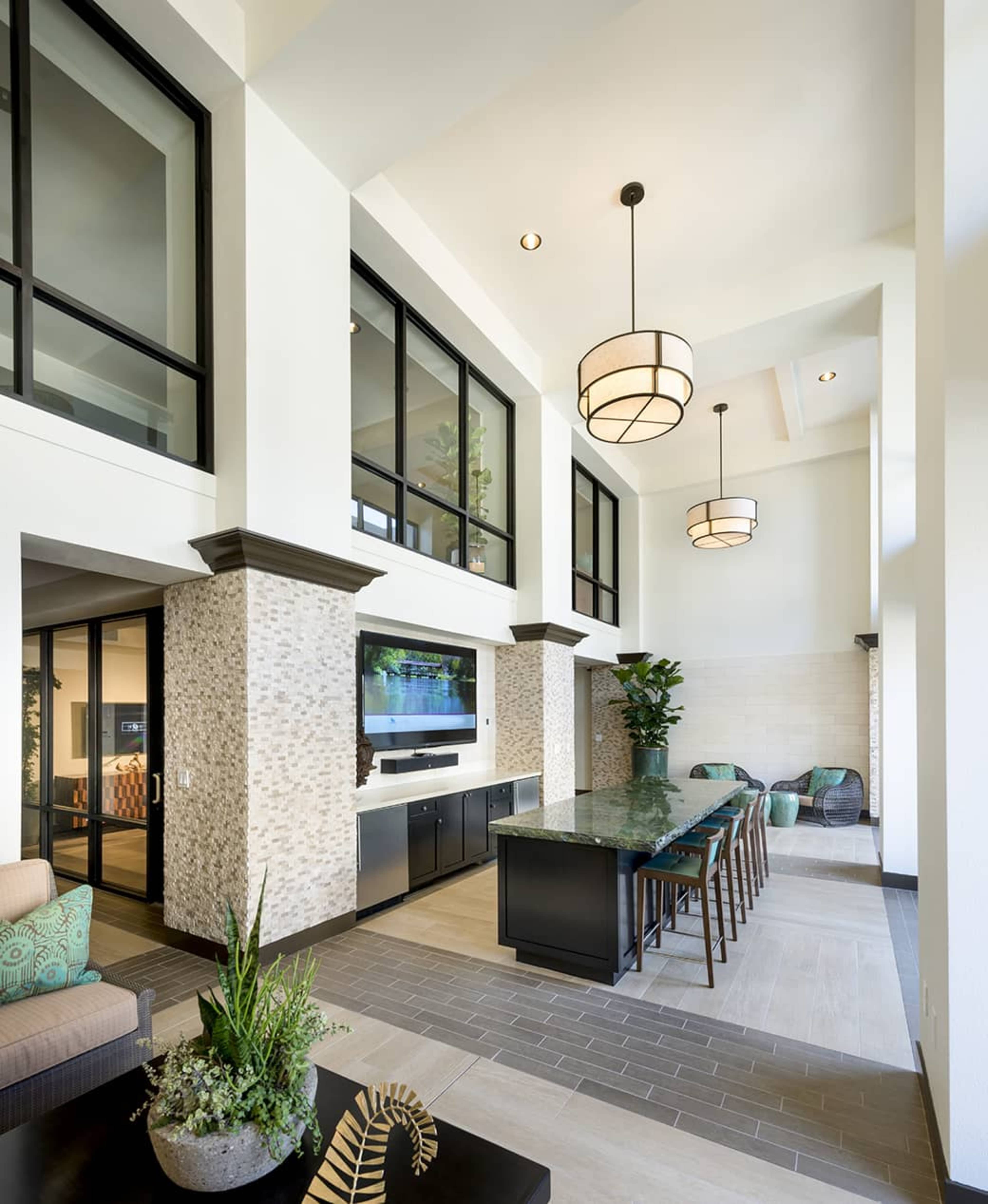 A modern communal area with a high ceiling, featuring a long table, contemporary lighting fixtures, and large windows.