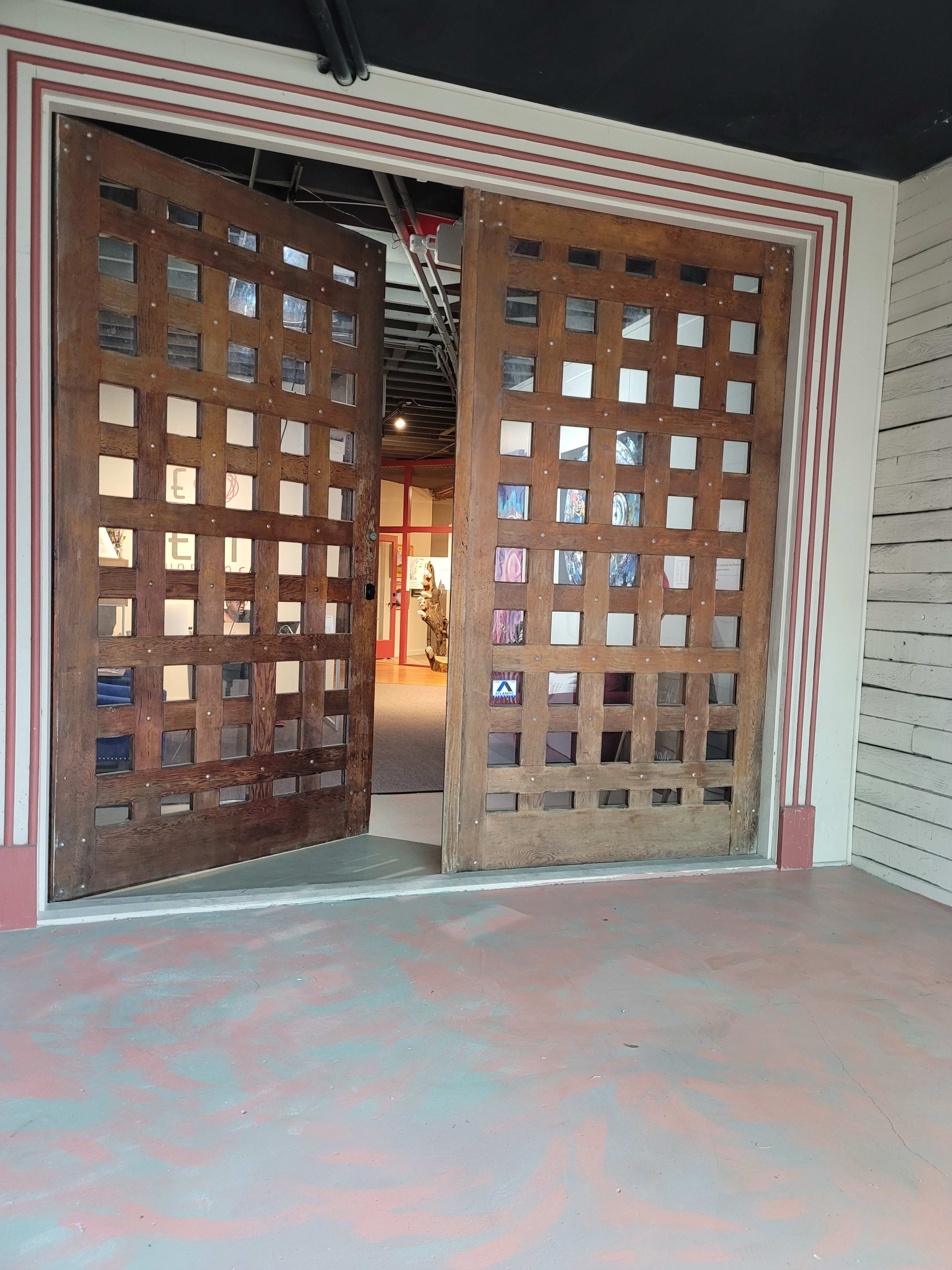 A set of large, wooden double doors with a grid pattern, partially opened to reveal a hallway and gallery beyond.