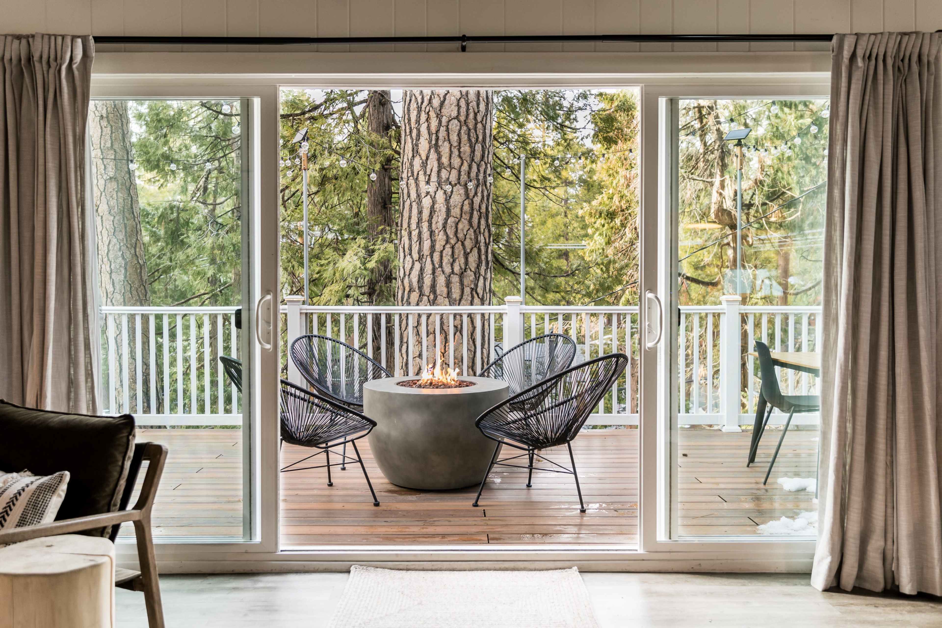 The image shows a cozy outdoor seating area with three black chairs arranged around a circular fire pit on a wooden deck surrounded by trees.