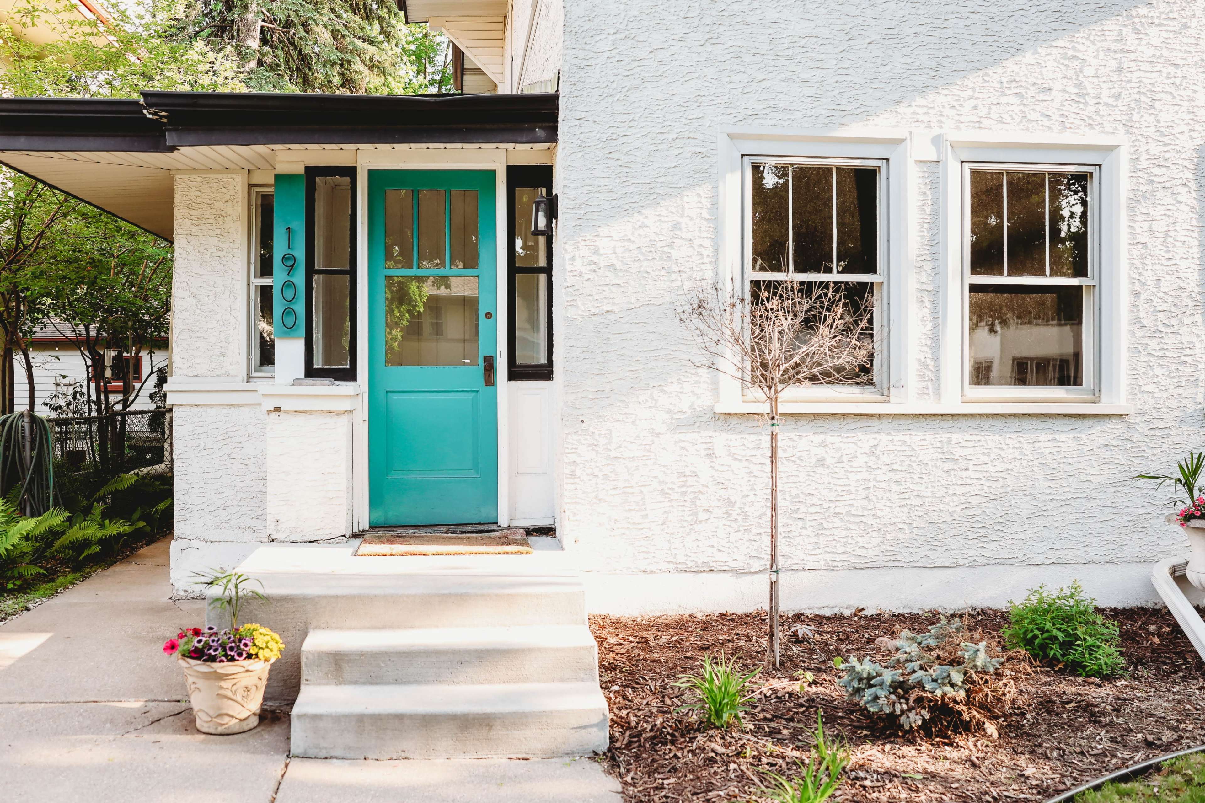 The image shows a house with a light-colored textured exterior, featuring a turquoise front door and landscaping that includes a small tree and various plants near the entryway.