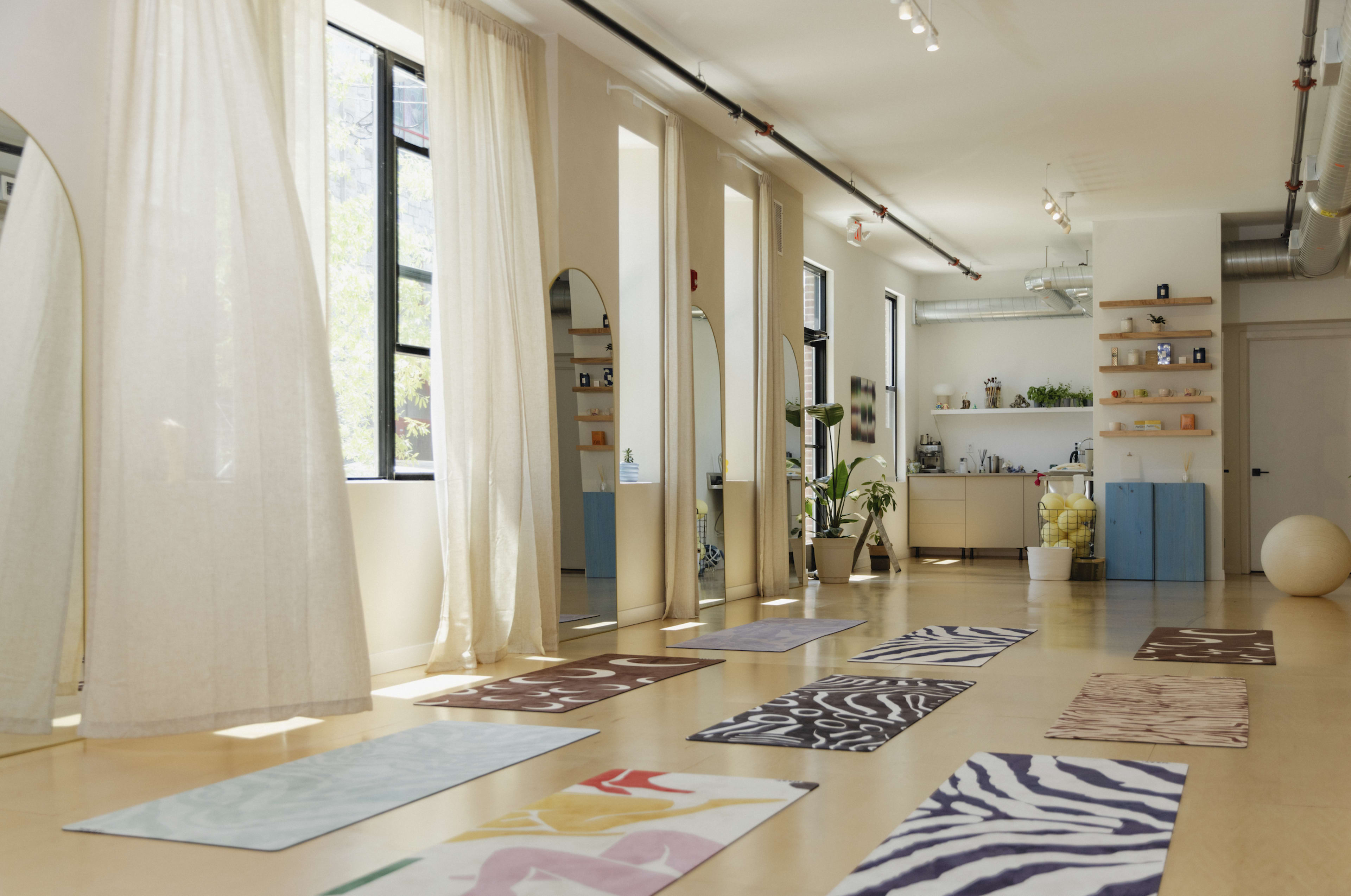 The image shows a bright, spacious yoga studio with several exercise mats arranged on the floor, large windows allowing natural light in, and a kitchenette in the background.