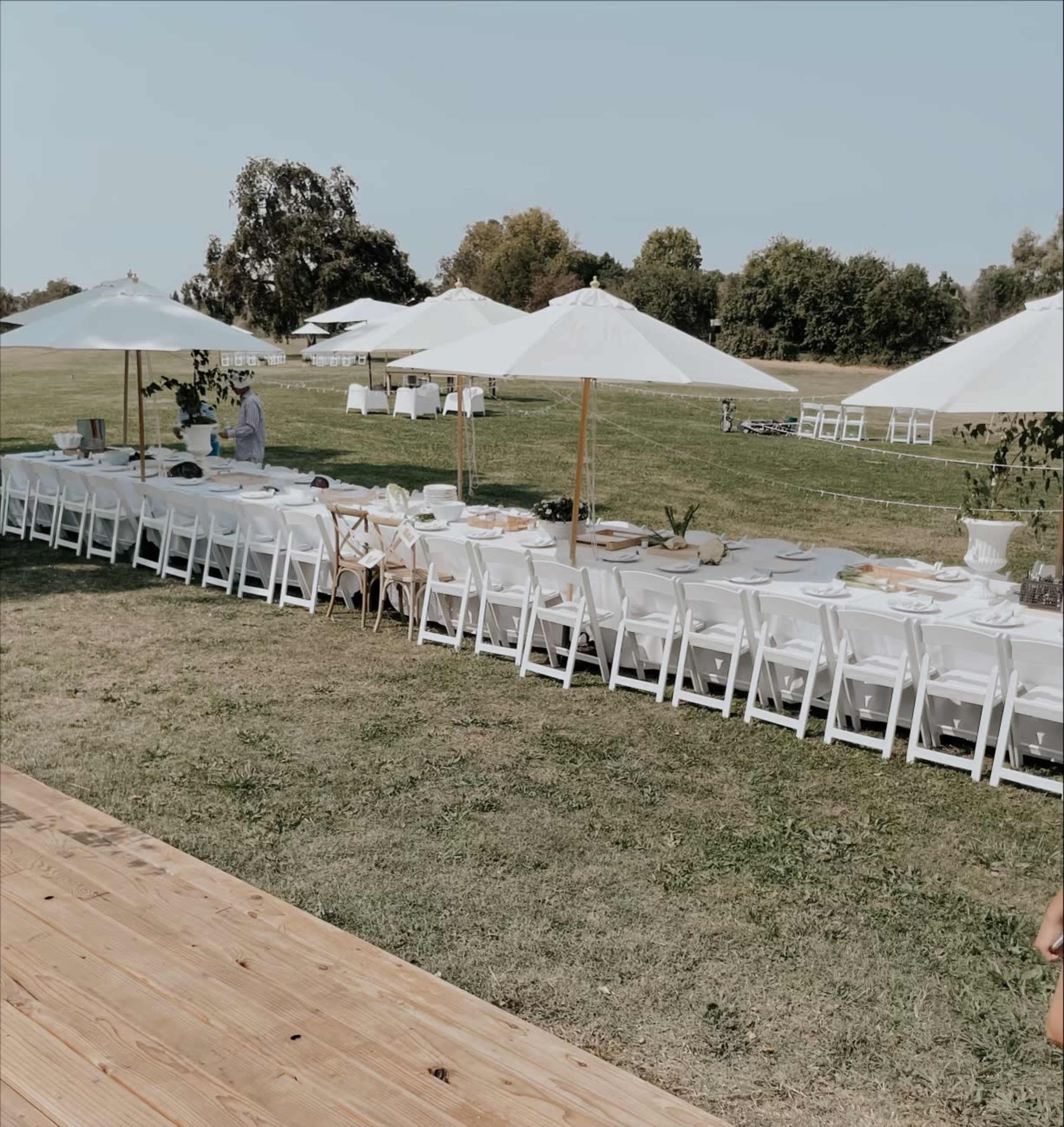 A long banquet table is set up outdoors under white umbrellas, surrounded by green grass and trees in the background.