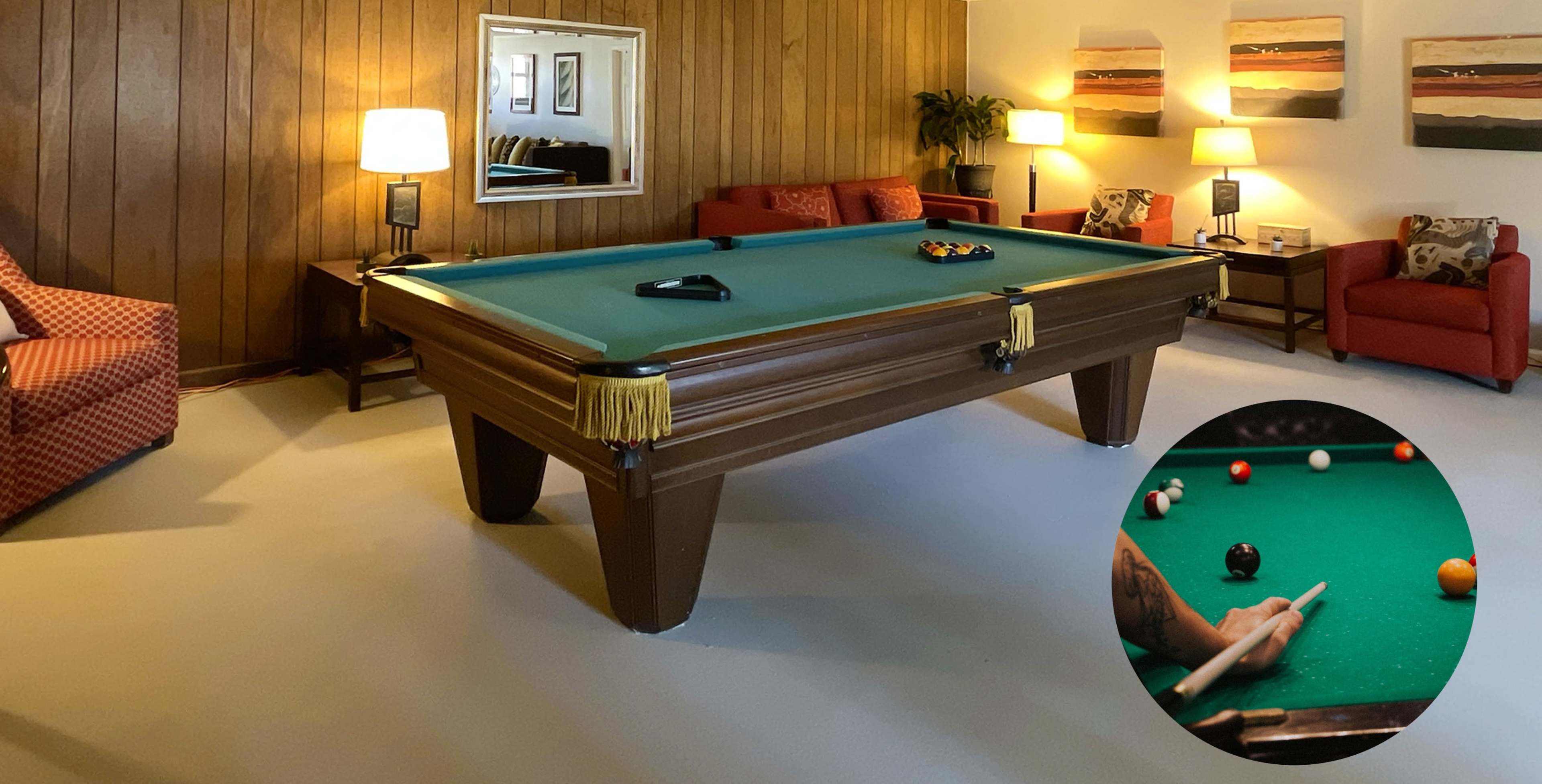 A game room featuring a green billiards table, two red armchairs, a mirror on the wall, and several framed artworks.