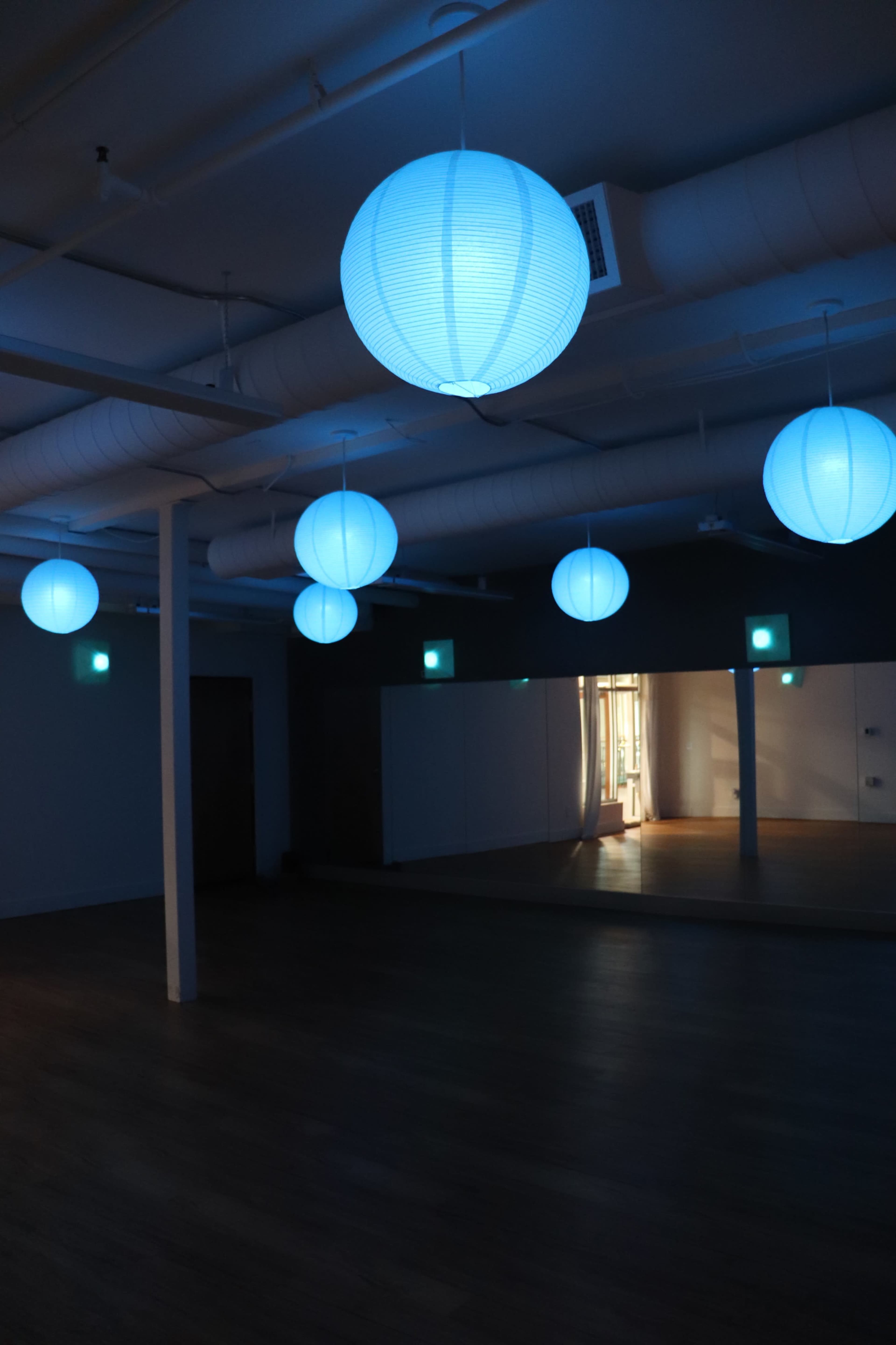 The image shows a spacious room with wooden floors, illuminated by several blue paper lanterns hanging from the ceiling and a large mirror on the wall.