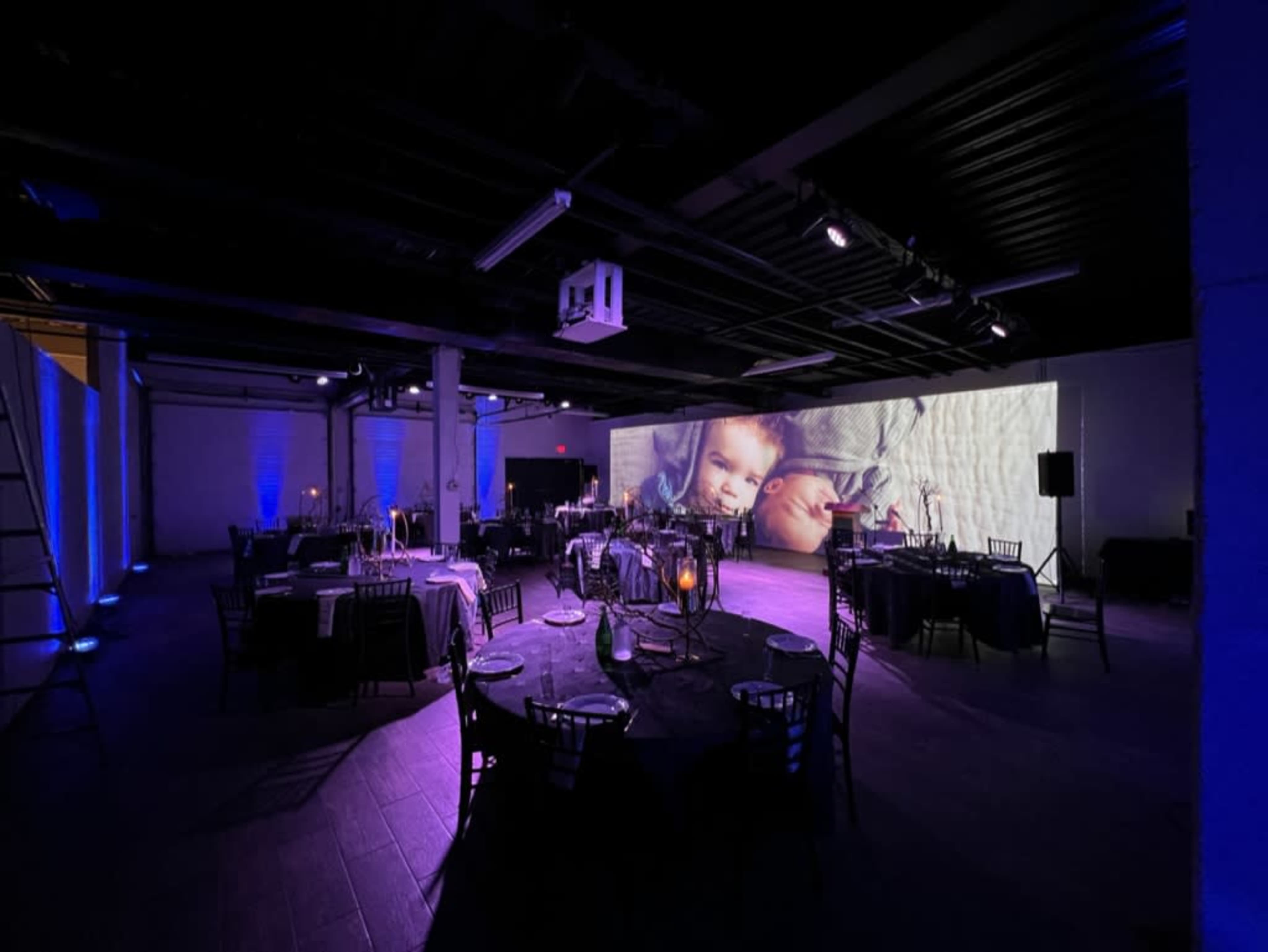 A large event space is set up with elegantly arranged tables, and a projection featuring two children on the wall.
