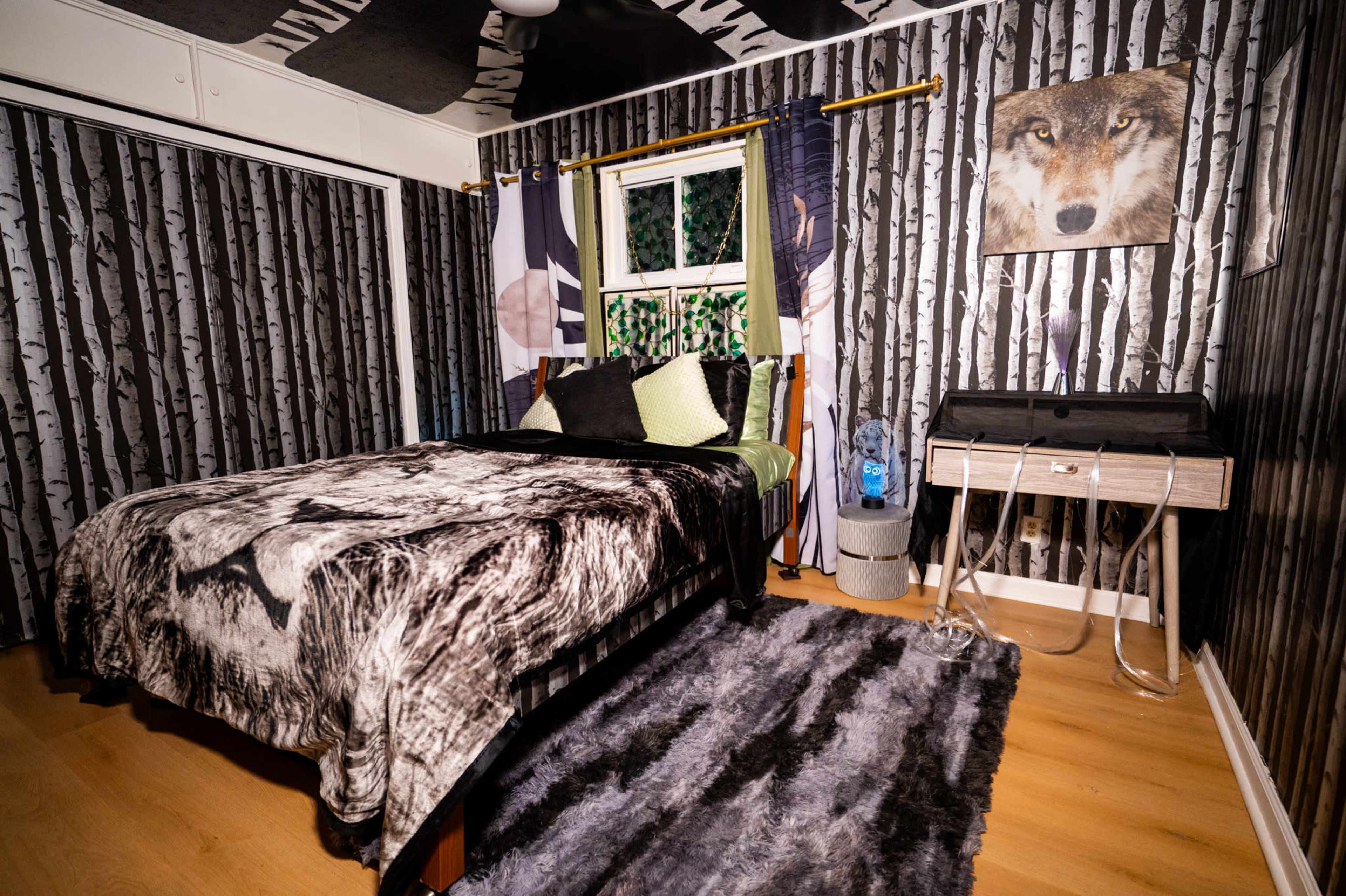 The image shows a bedroom with a bed covered in a wolf-themed blanket, black and white birch tree wallpaper, and a window with green foliage visible outside.