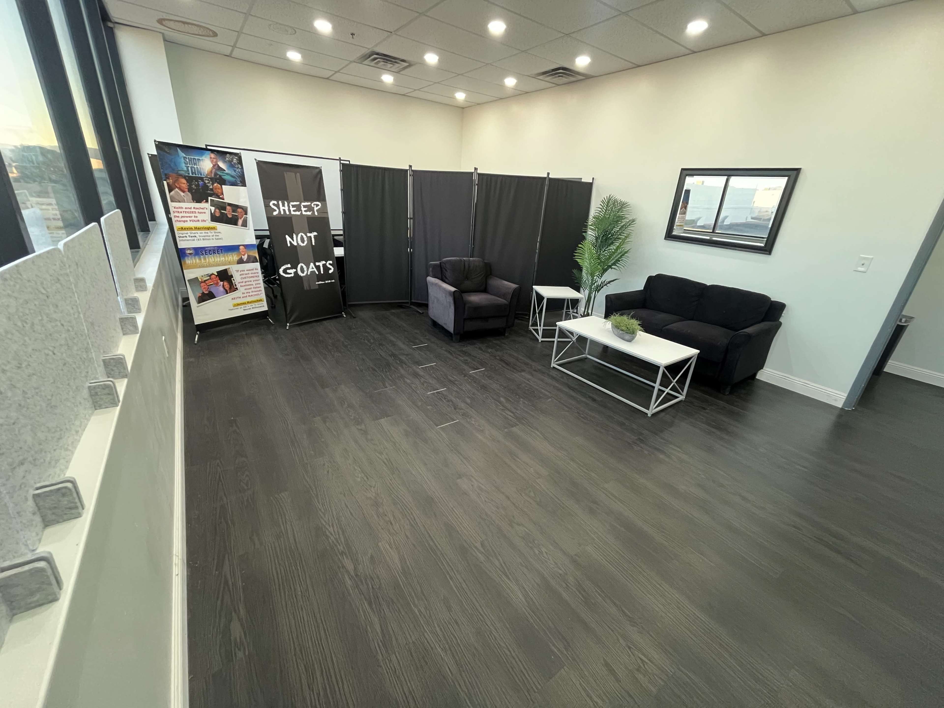 The image shows a spacious waiting area featuring two black chairs, a coffee table, a potted plant, and a backdrop with the text “SHEEP NOT GOATS” along with promotional materials.
