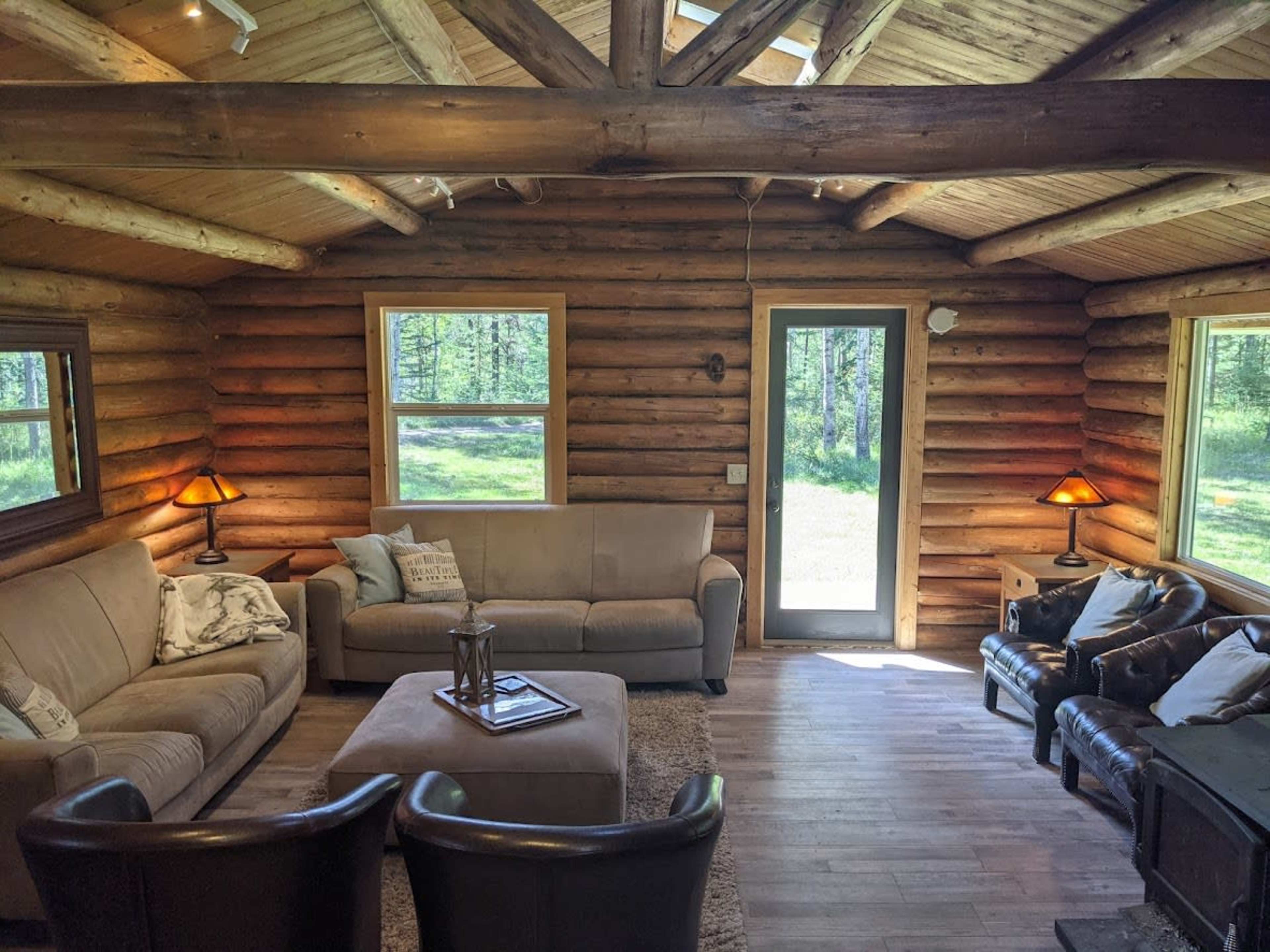 The interior of a log cabin living room features a beige sectional sofa, a circular coffee table, two dark armchairs, and large windows providing a view of the outdoor greenery.