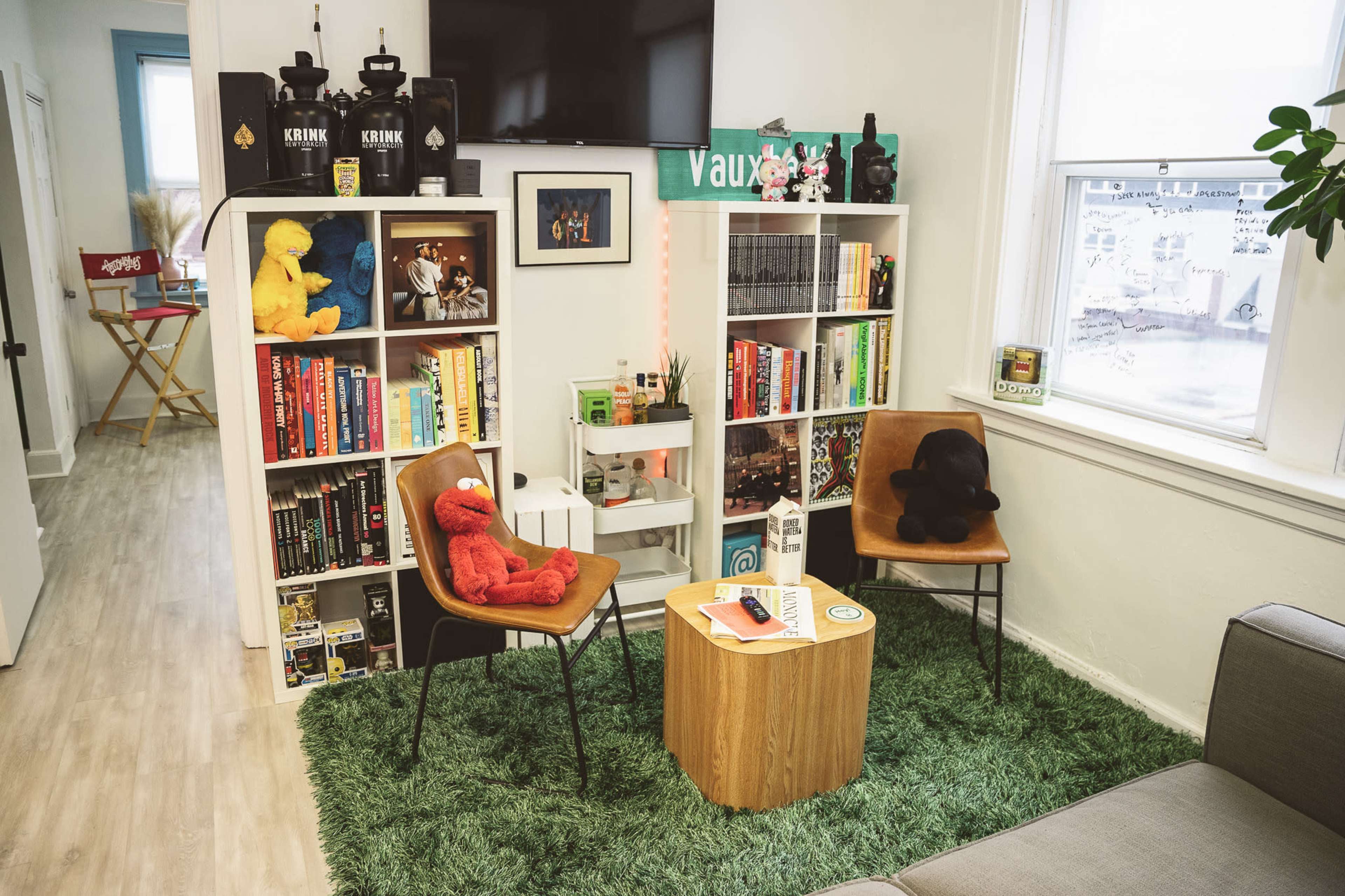 A cozy reading nook with two chairs, a round wooden table, bookshelves filled with books and collectibles, and greenery on the floor.