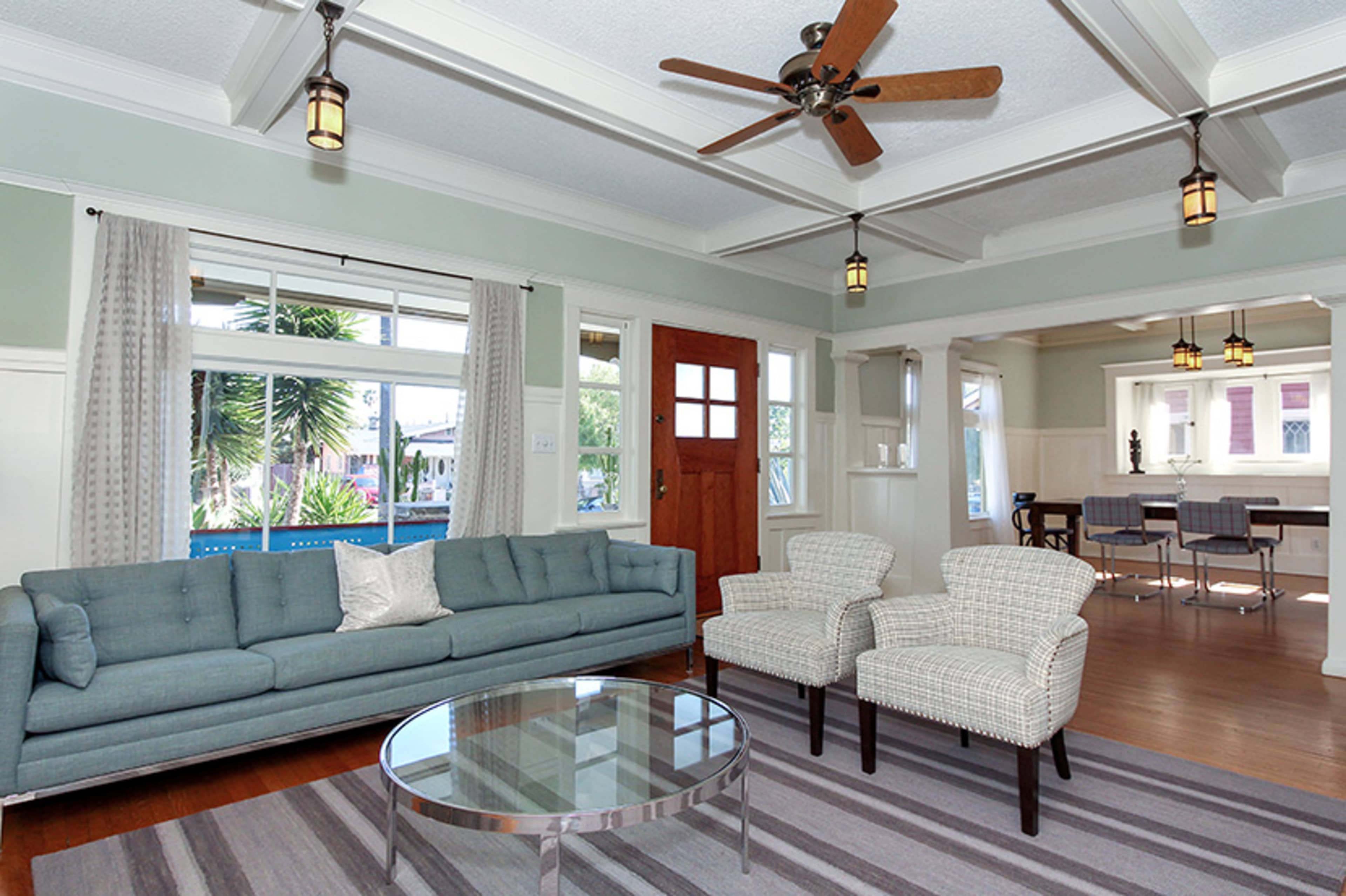 The room features a light blue sofa, two patterned chairs, a round glass coffee table, and large windows that allow natural light to fill the space.