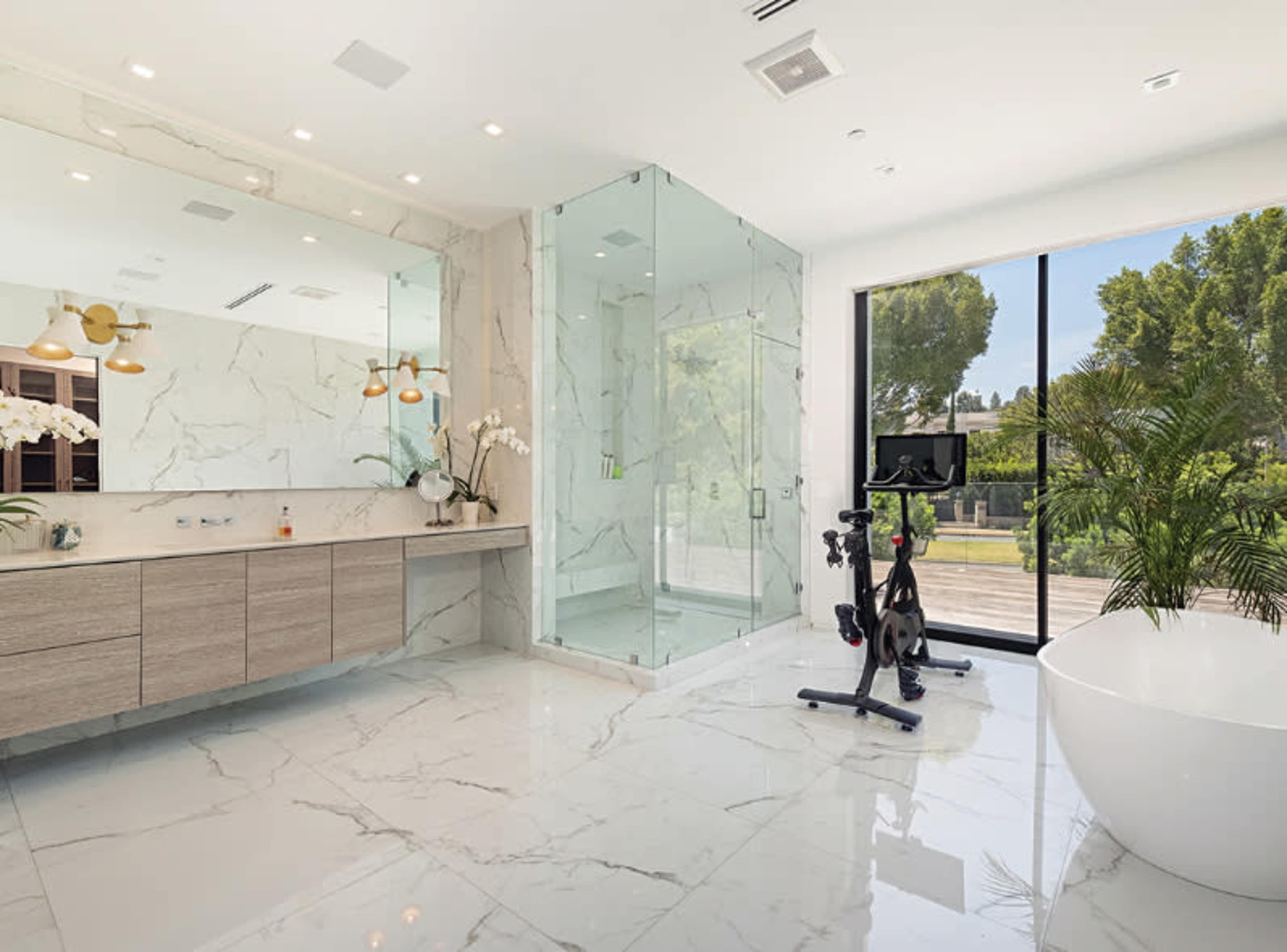 A modern bathroom featuring a sleek glass shower, a freestanding bathtub, and a workout bike, all set against a backdrop of marble flooring and large windows.