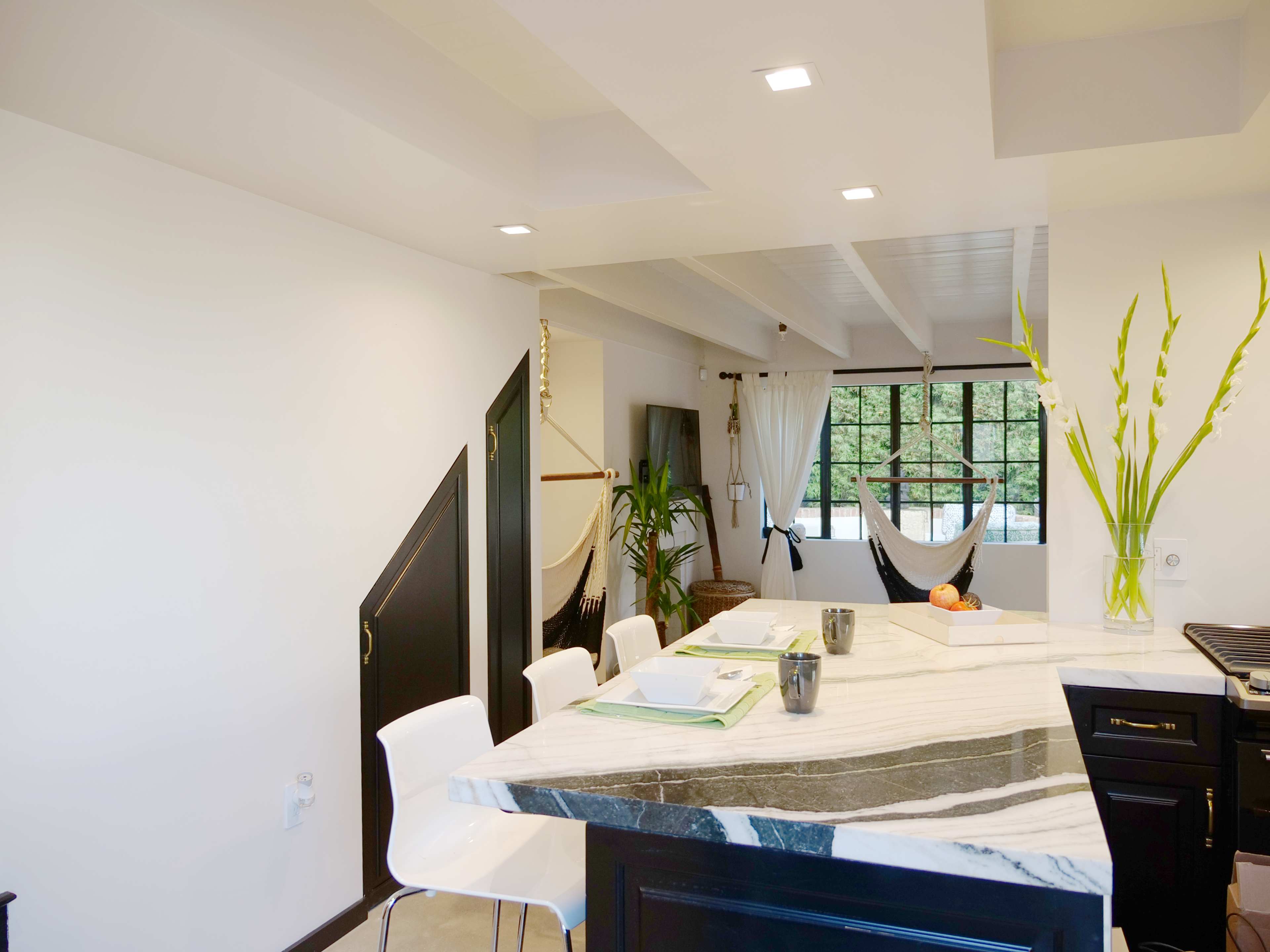 A modern kitchen with a marble countertop, white chairs around a dining area, and a hammock visible in the background.