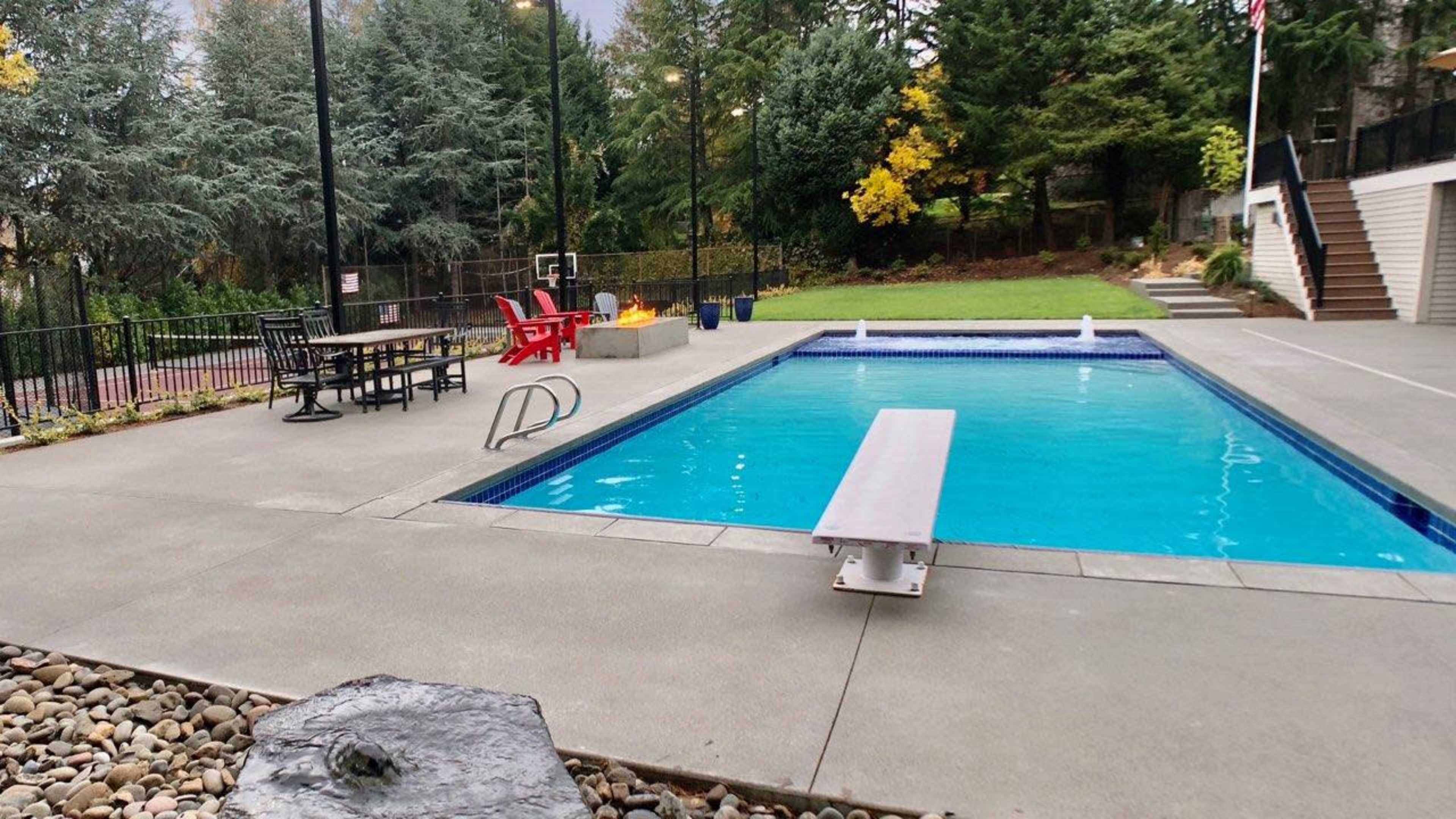 The image shows a swimming pool surrounded by a patio area with a diving board, lounge chairs, and trees in the background.