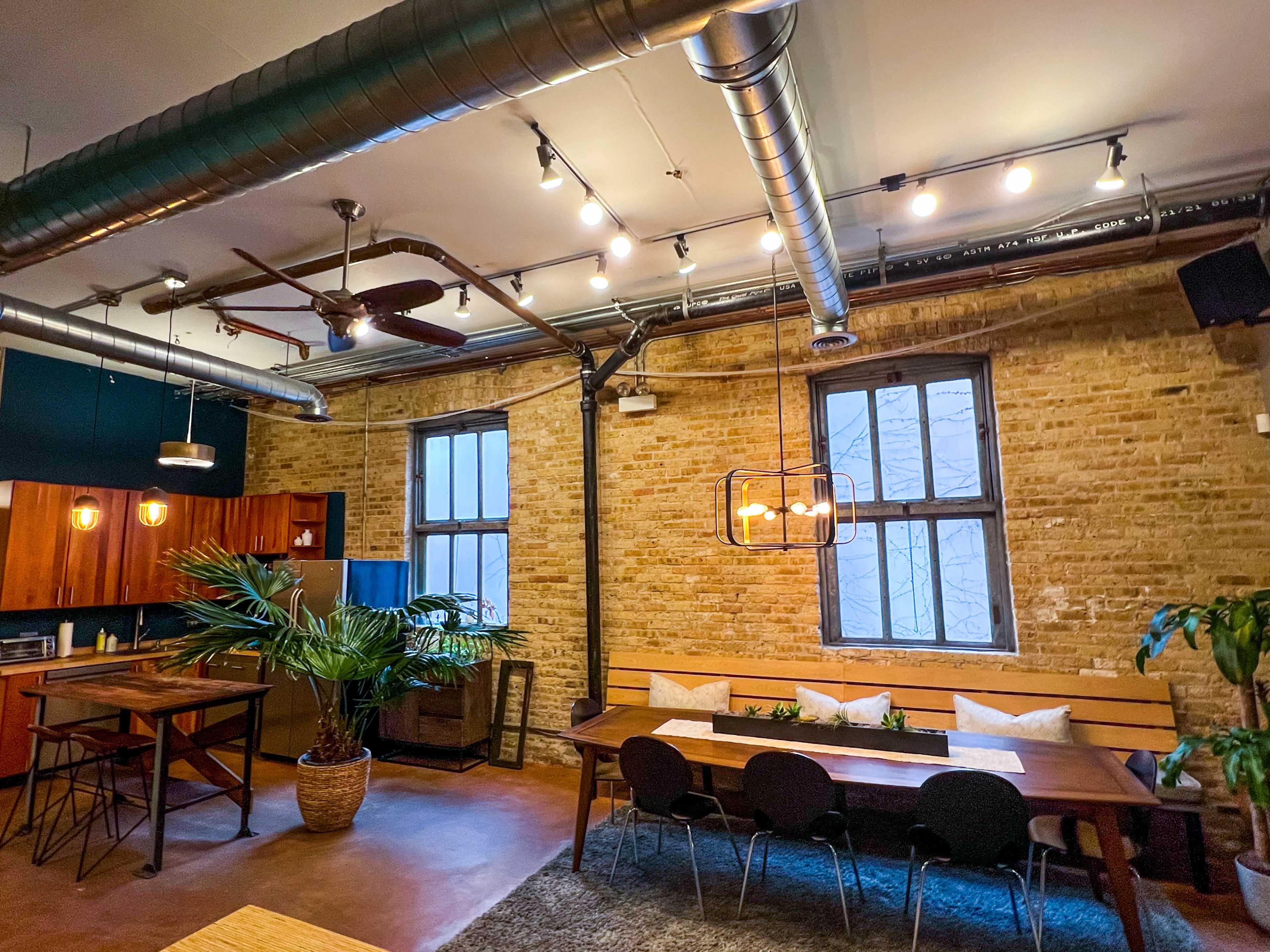 The image shows a spacious, modern kitchen and dining area with exposed brick walls, industrial lighting, and wooden furniture.