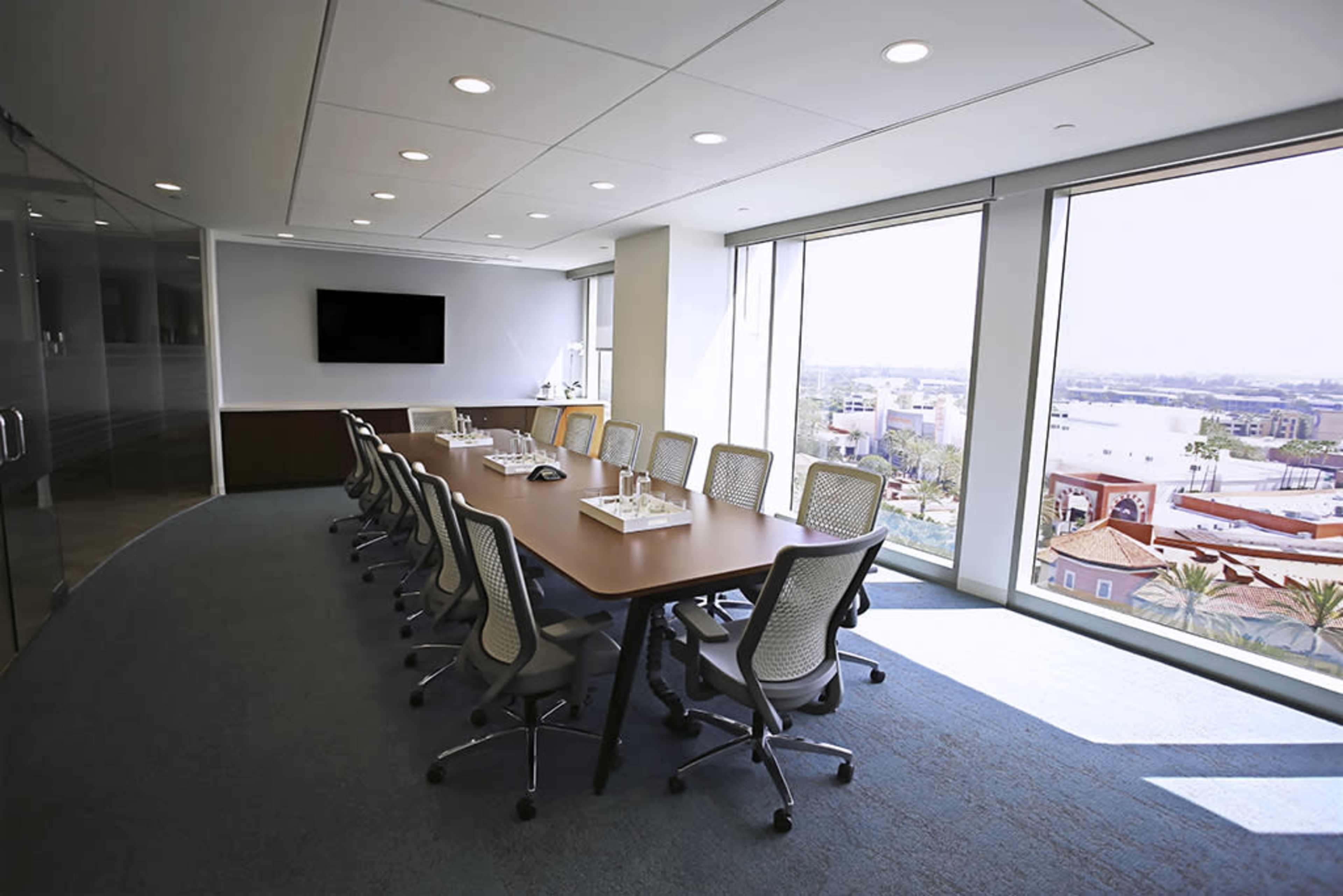 A large conference room with a rectangular table, eight ergonomic chairs, and a scenic view of a waterfront through large windows.