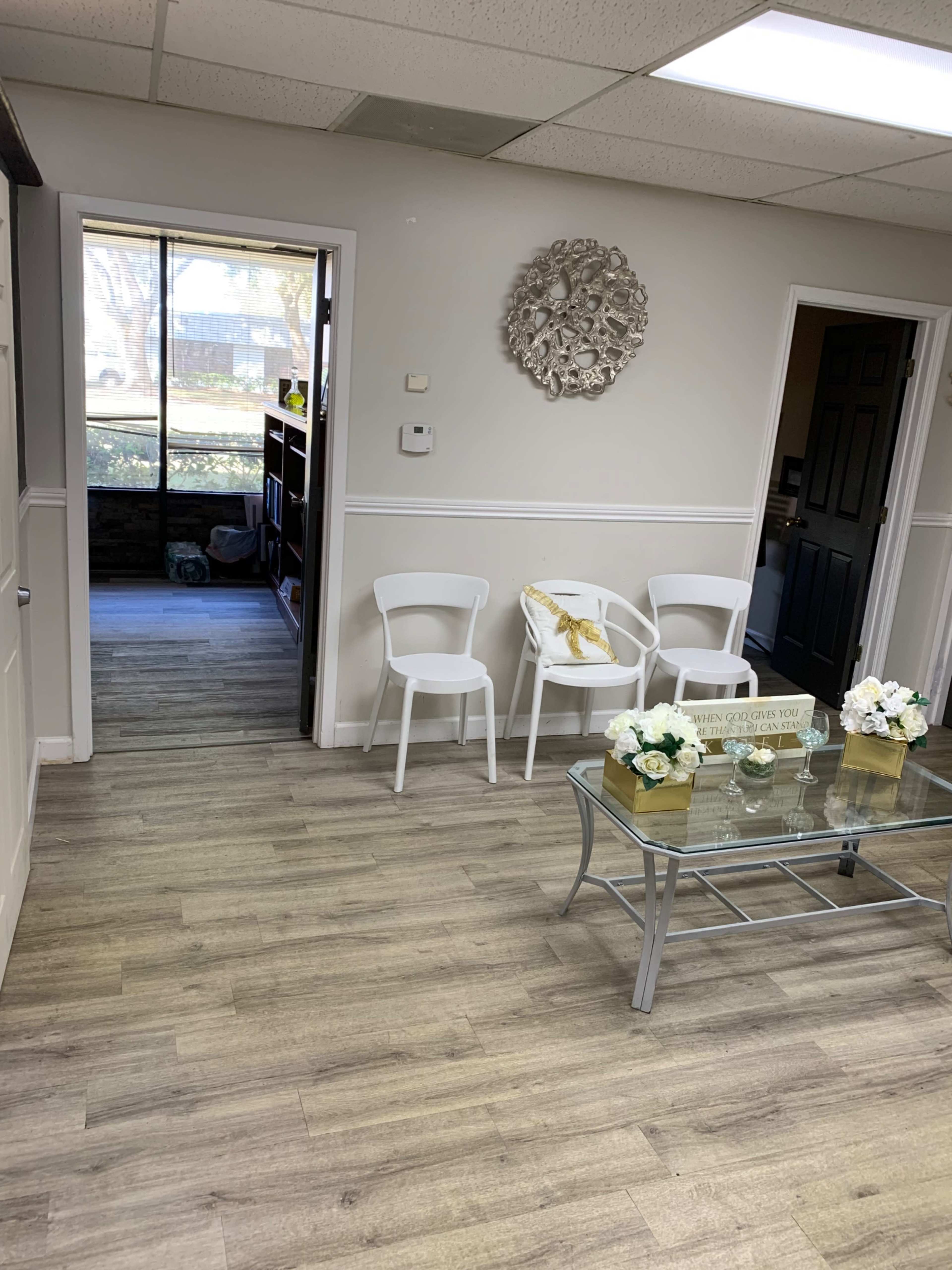 The image shows a waiting area with white chairs, a glass coffee table with decorative items, and a doorway leading to another room.