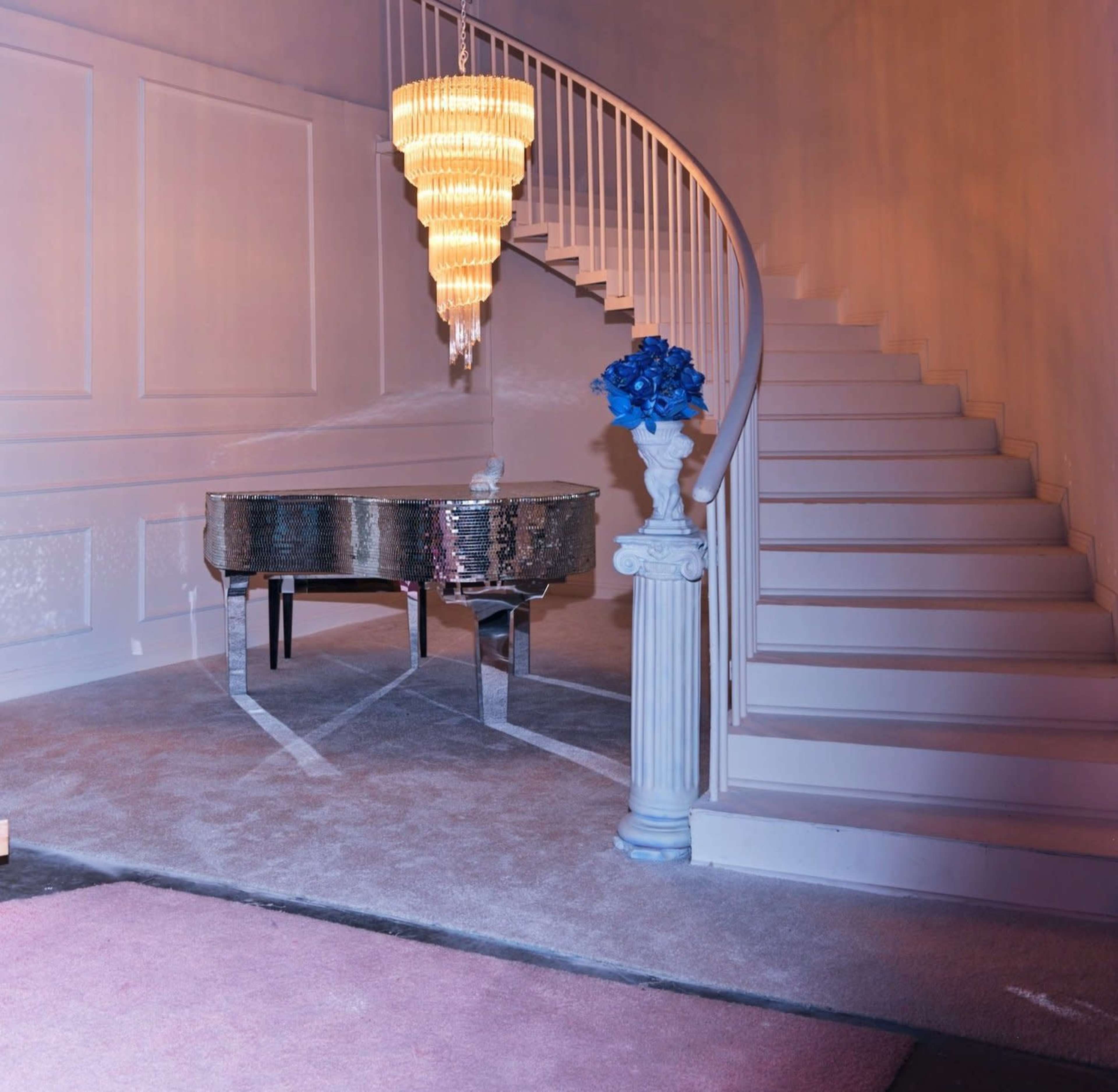 A spiral staircase is illuminated beside a reflective grand piano, with a vase of blue flowers on a column nearby.