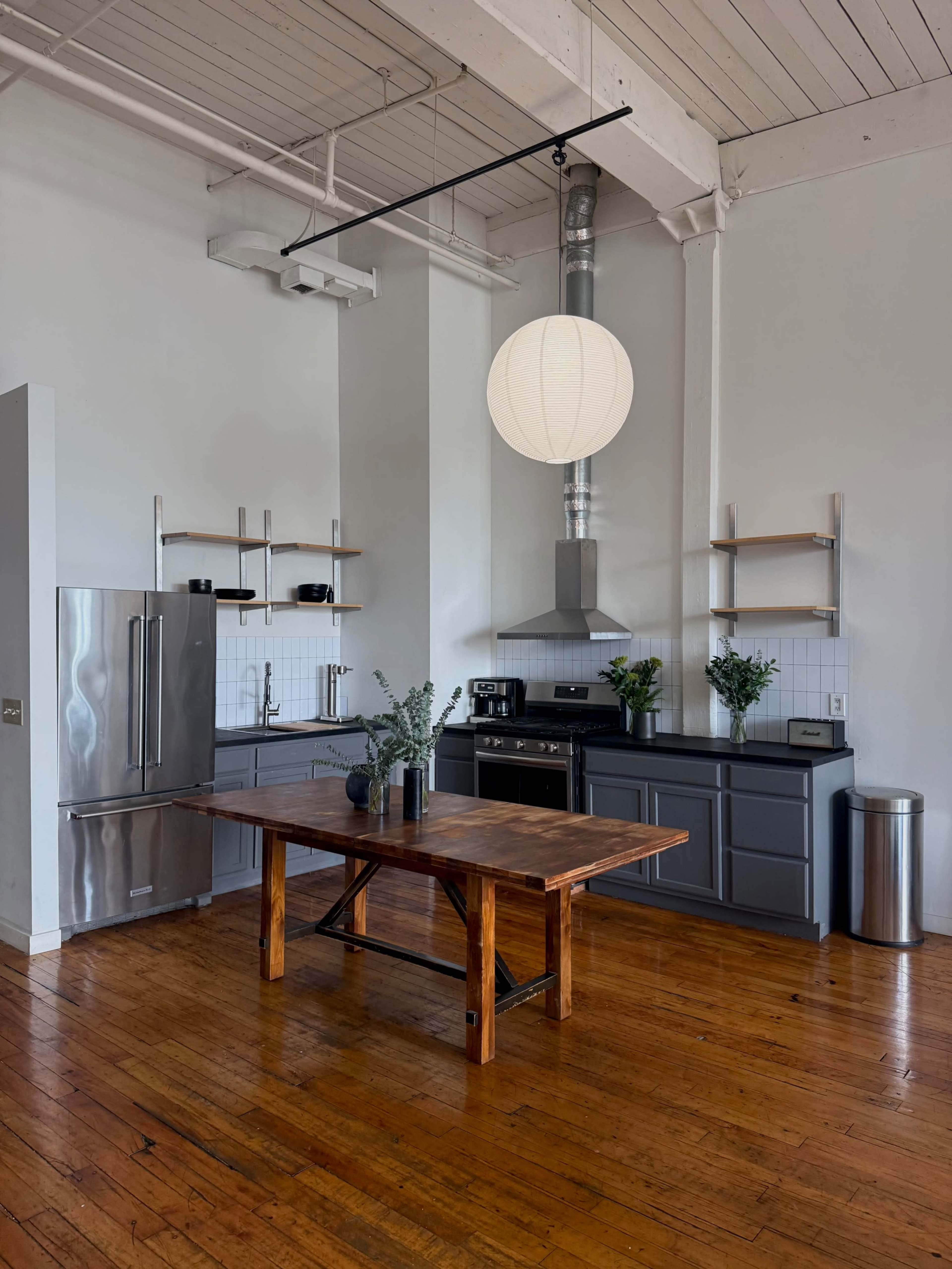 The image shows a modern kitchen featuring stainless steel appliances, wooden flooring, and a central dining table surrounded by shelves and plants.