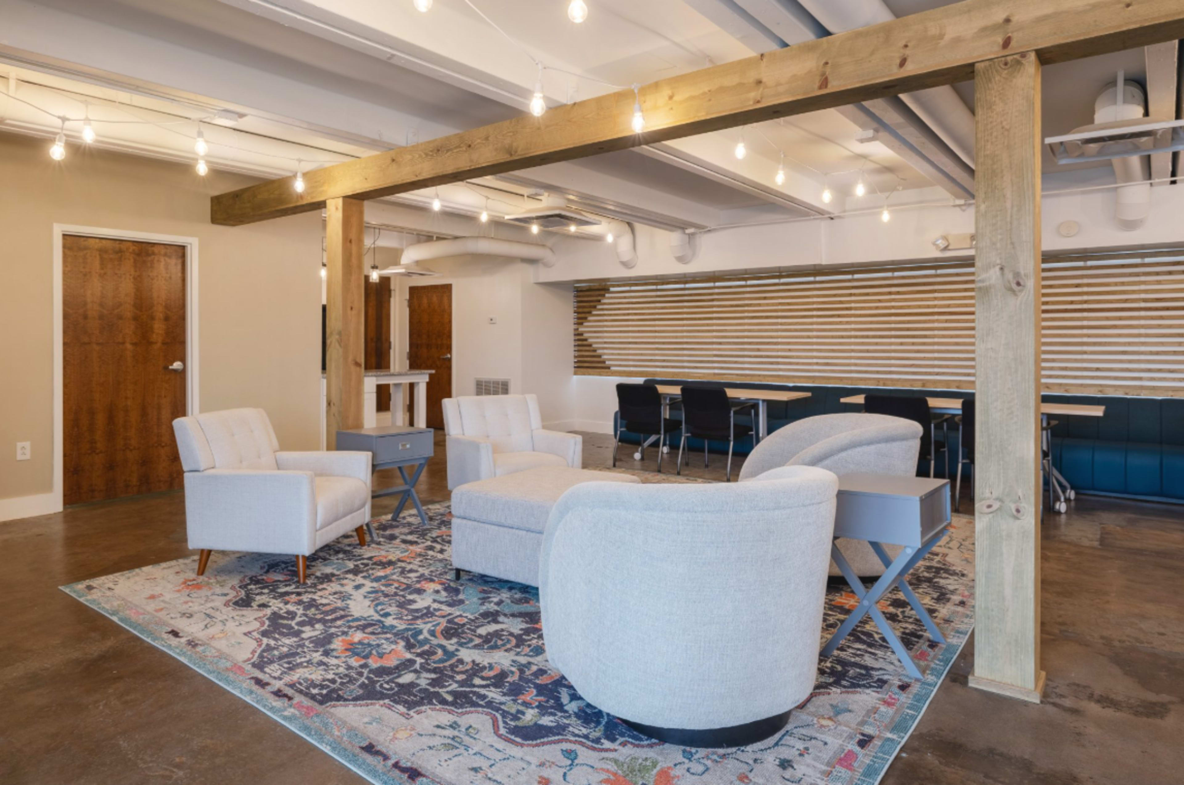 The image shows a modern, open lounge area featuring a gray upholstered seating arrangement on a decorative rug, with wooden beams overhead and a conference area in the background.