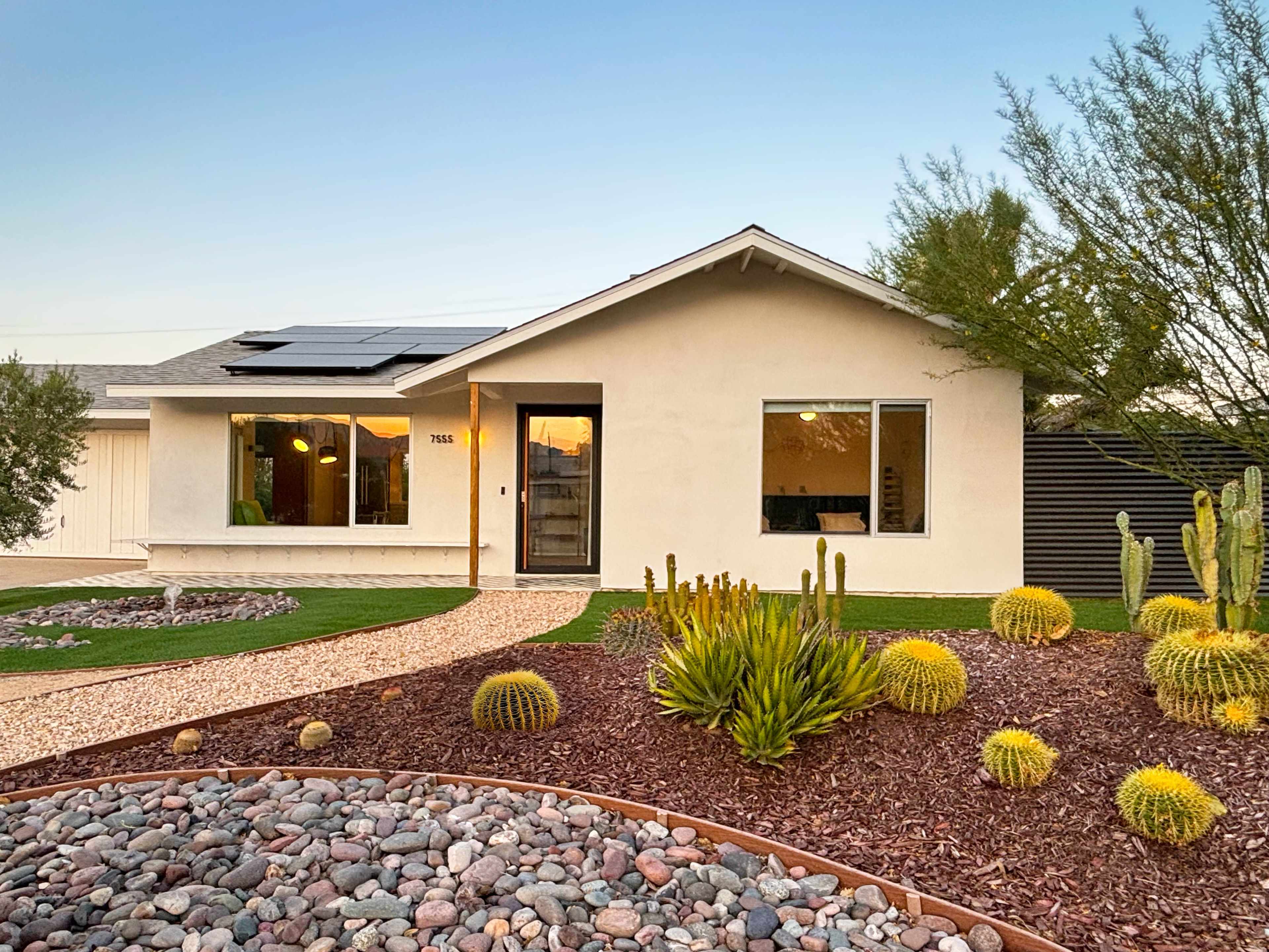 The image shows a modern single-story house with a solar panel on the roof, surrounded by landscaped gardens featuring rocks, cacti, and shrubs.