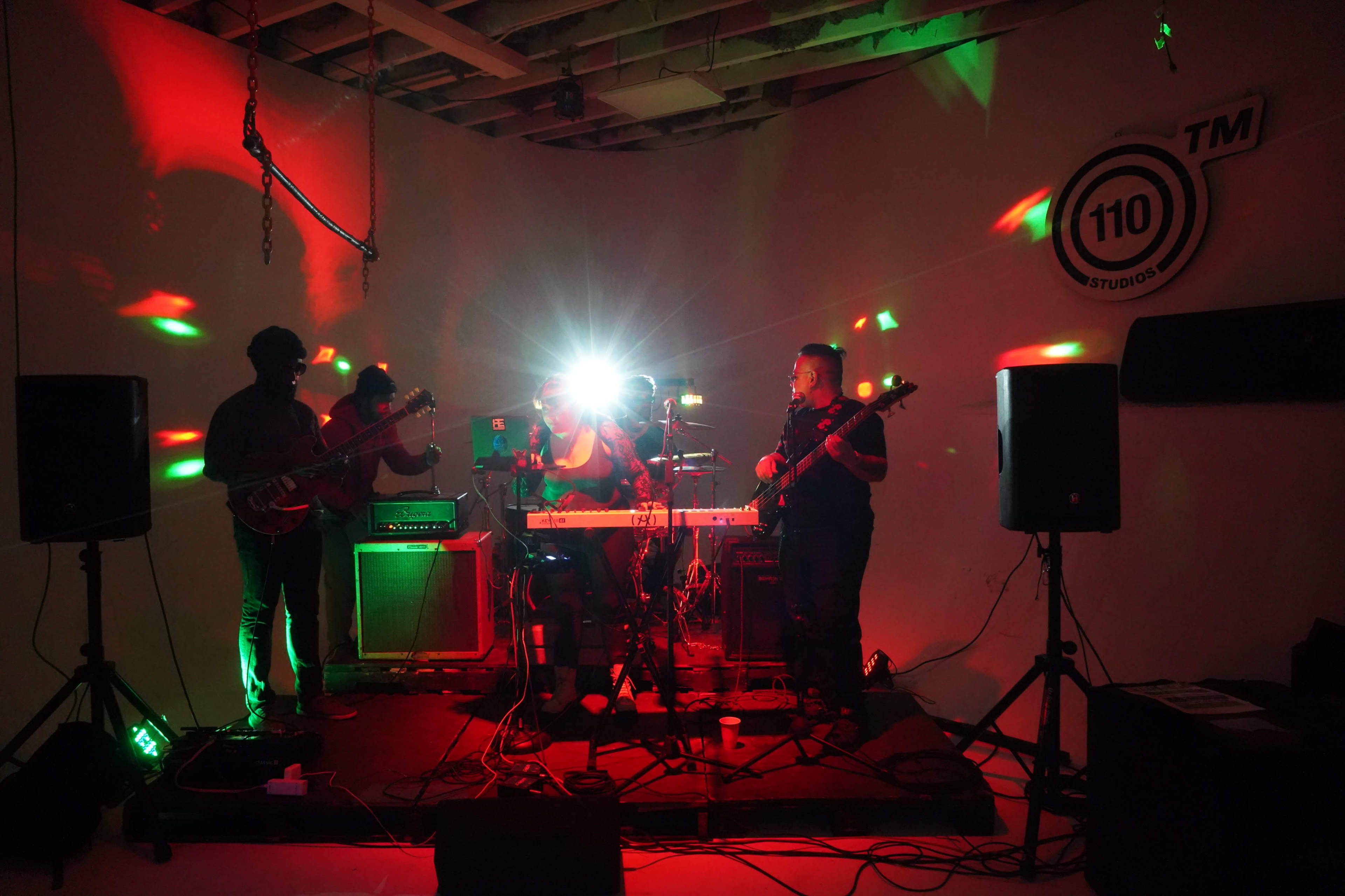 A band performs on stage with colorful lights illuminating the space, surrounded by amplifiers and musical instruments.