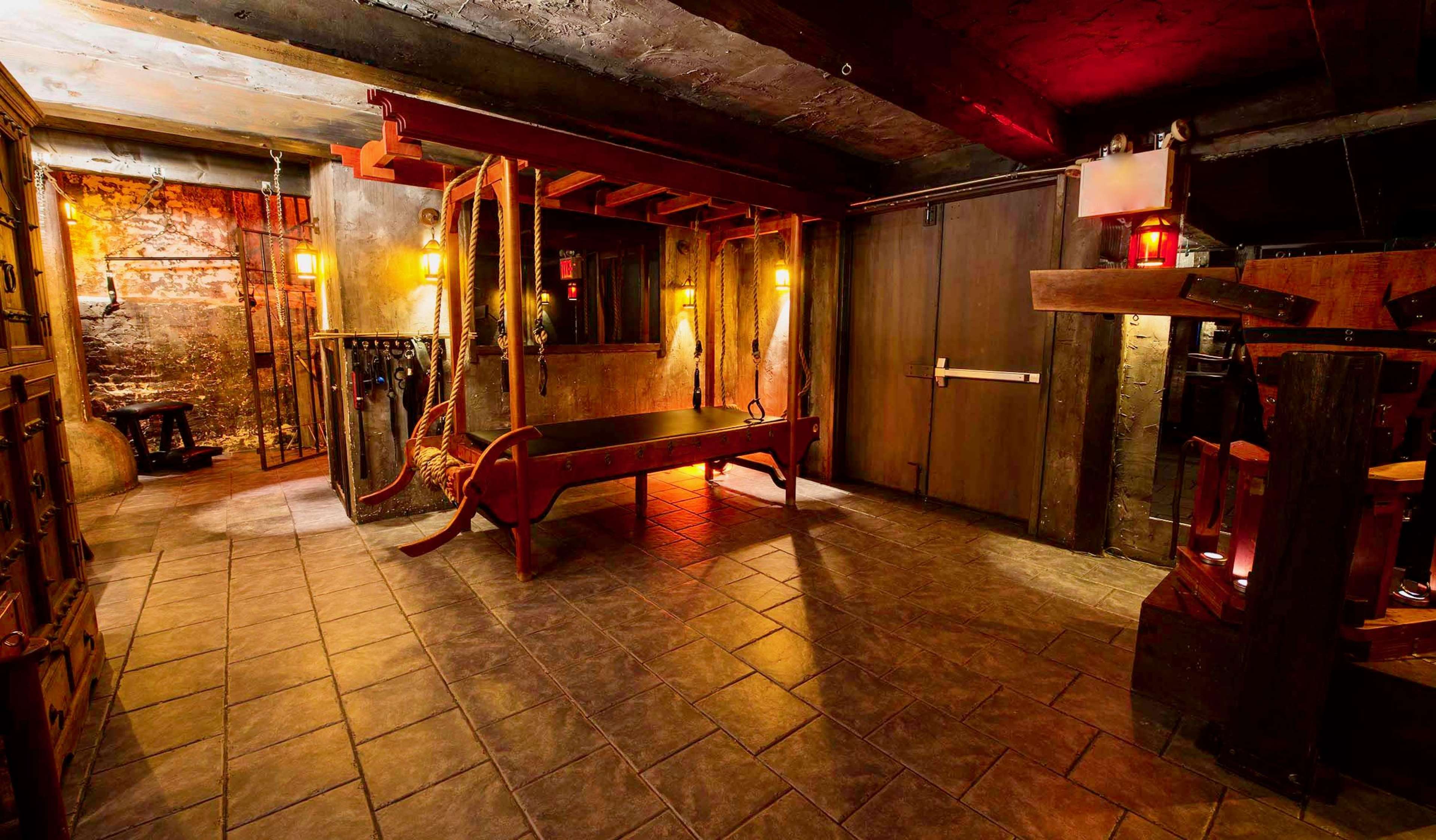 The image shows a dimly lit underground room with stone walls, featuring a wooden torture device, chains, and several lanterns illuminating the space.