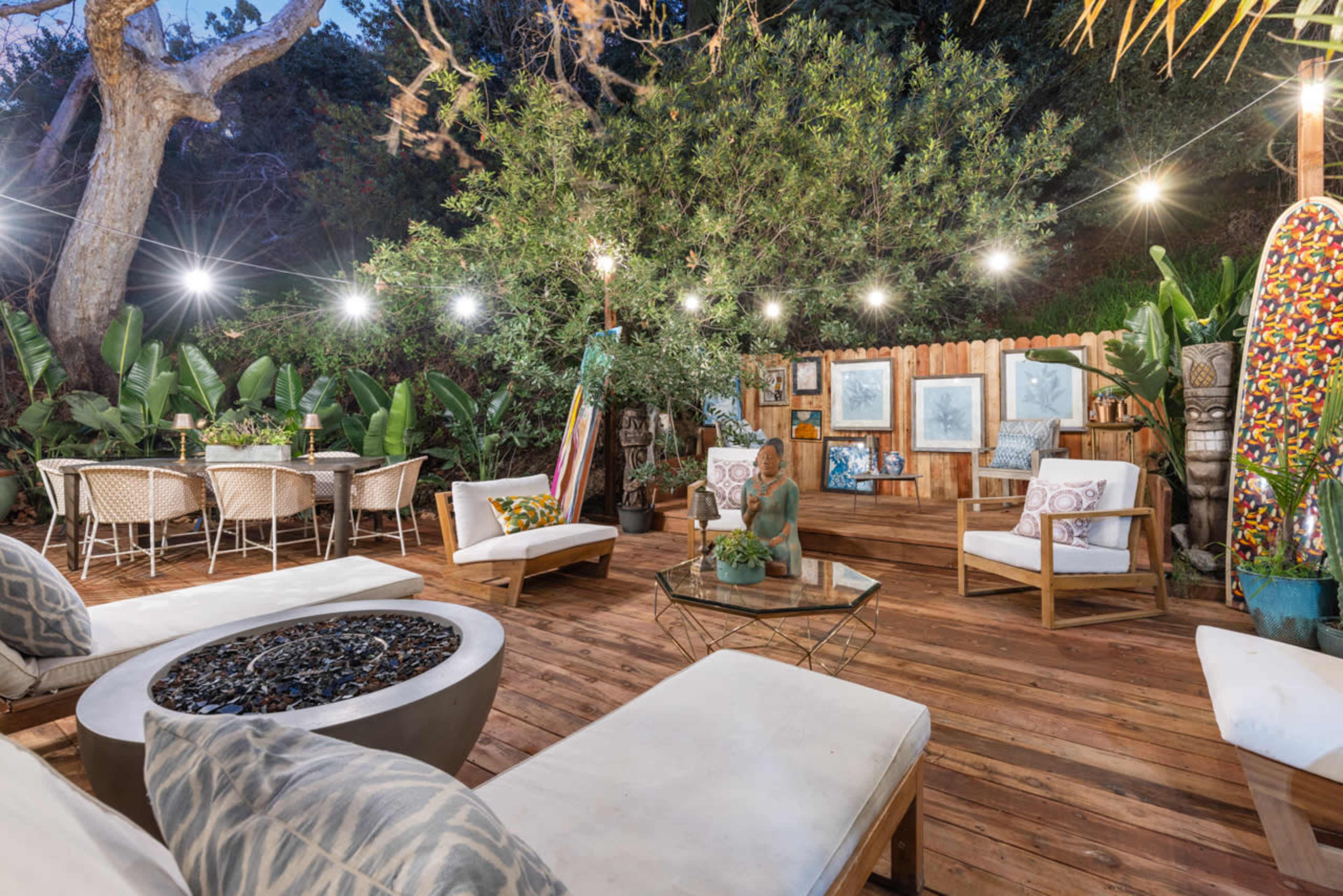 The image shows a cozy outdoor patio area with wooden flooring, various seating arrangements, a dining table, and decorative plants, illuminated by string lights.